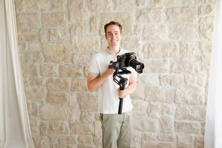 Ben holding a camera smiling in front of a stone wall