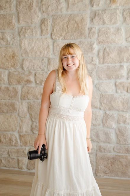 Amelia in a white dress holding a camera against a stone wall