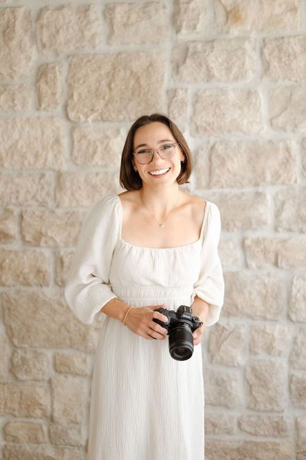 Tyler in a white dress holding a camera against a stone wall.
