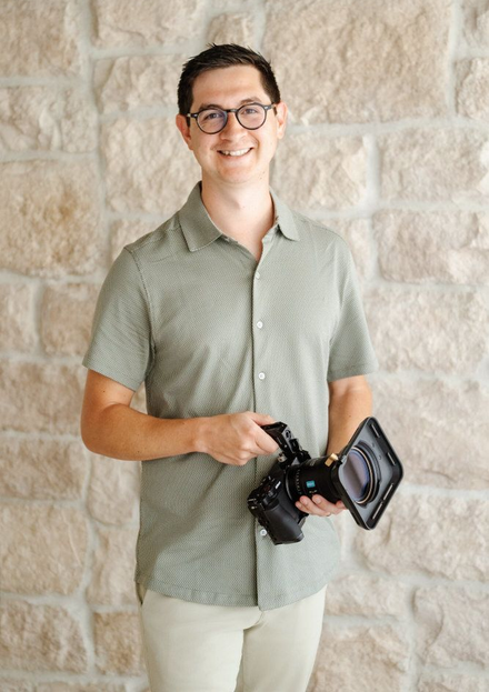 Dan holding a camera standing against a stone wall and smiling.