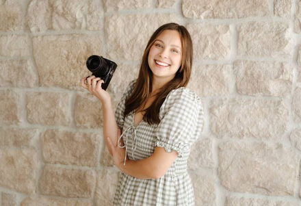 Emma holding a camera standing against a stone wall and smiling.