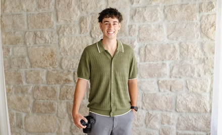 Parker in a green shirt and gray pants holding a camera, standing against a stone wall.