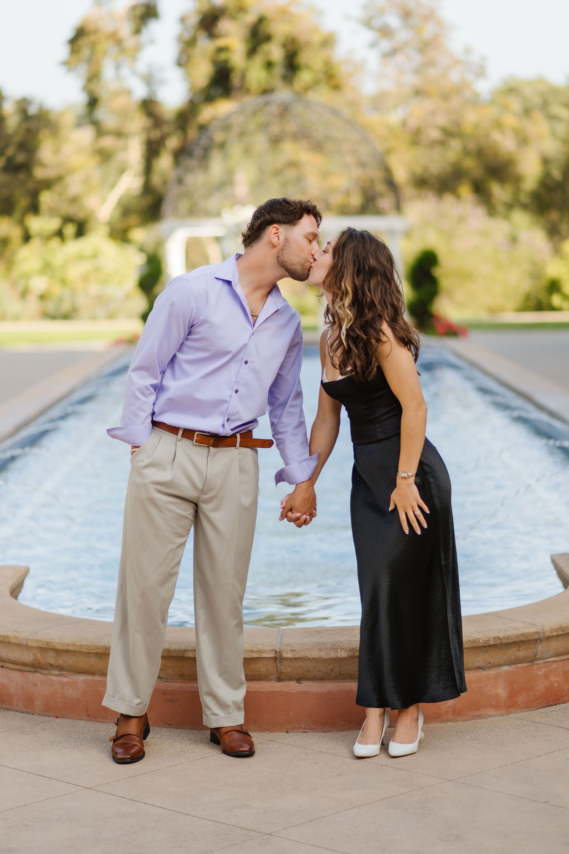 A couple leaning in for a kiss by a fountain, with greenery in the background at a proposal with The Yes Girls