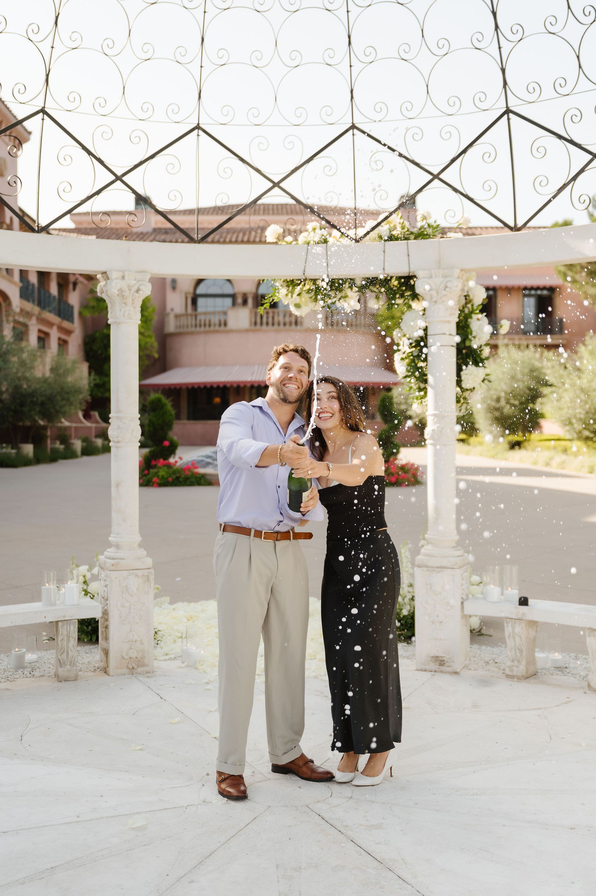 Couple standing under a white arch outdoors, smiling and releasing confetti at a proposal with The Yes Girls