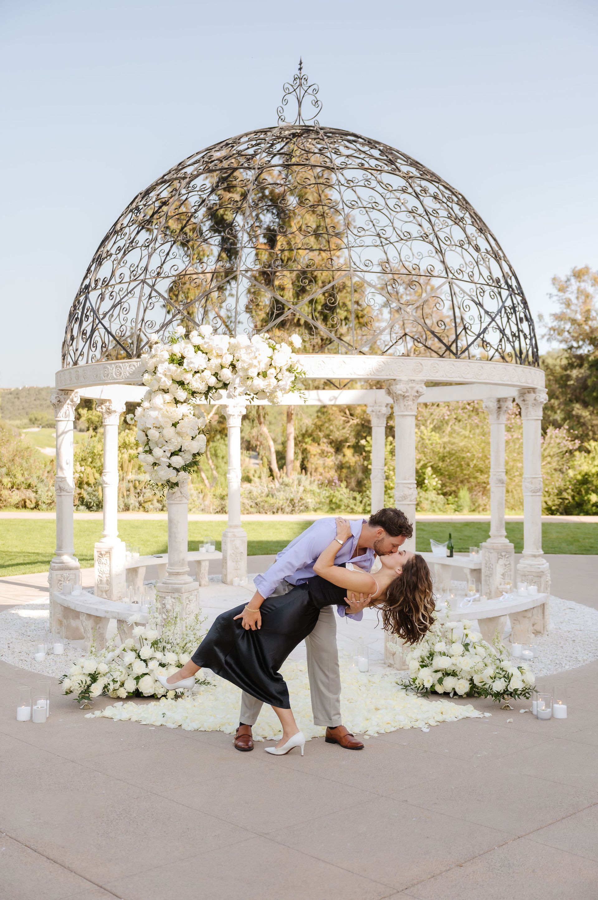 Couple kissing under a white floral gazebo in a garden setting for a proposal with The Yes Girls
