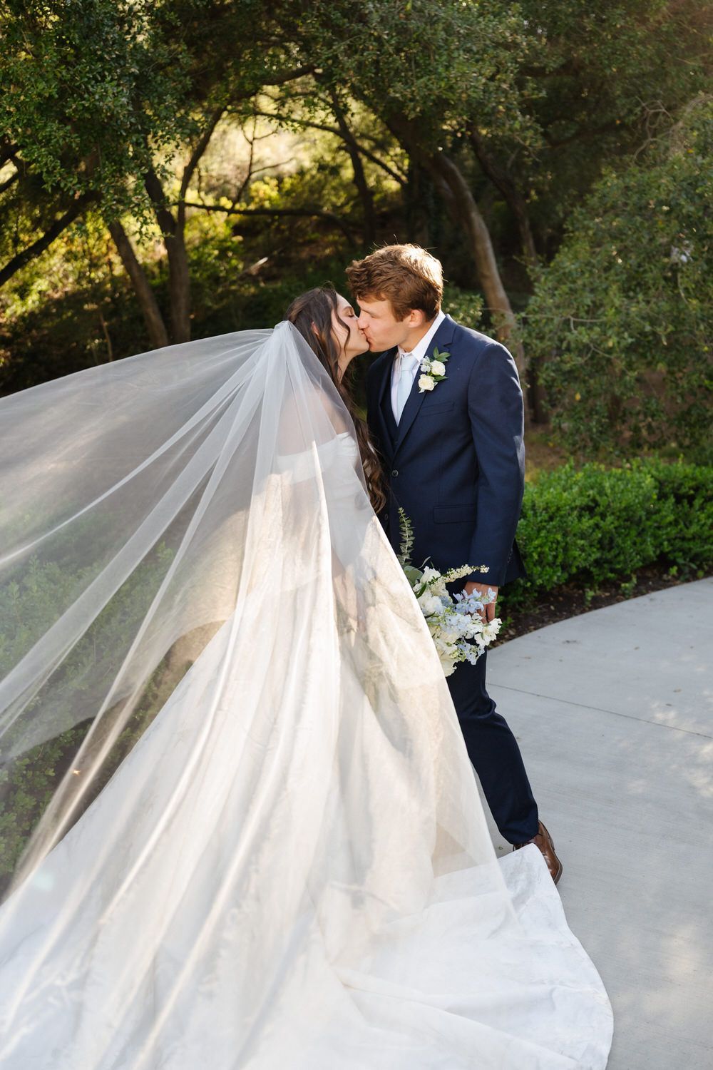 Bride and groom kiss outdoors on a garden path, bride’s veil flowing in the breeze.