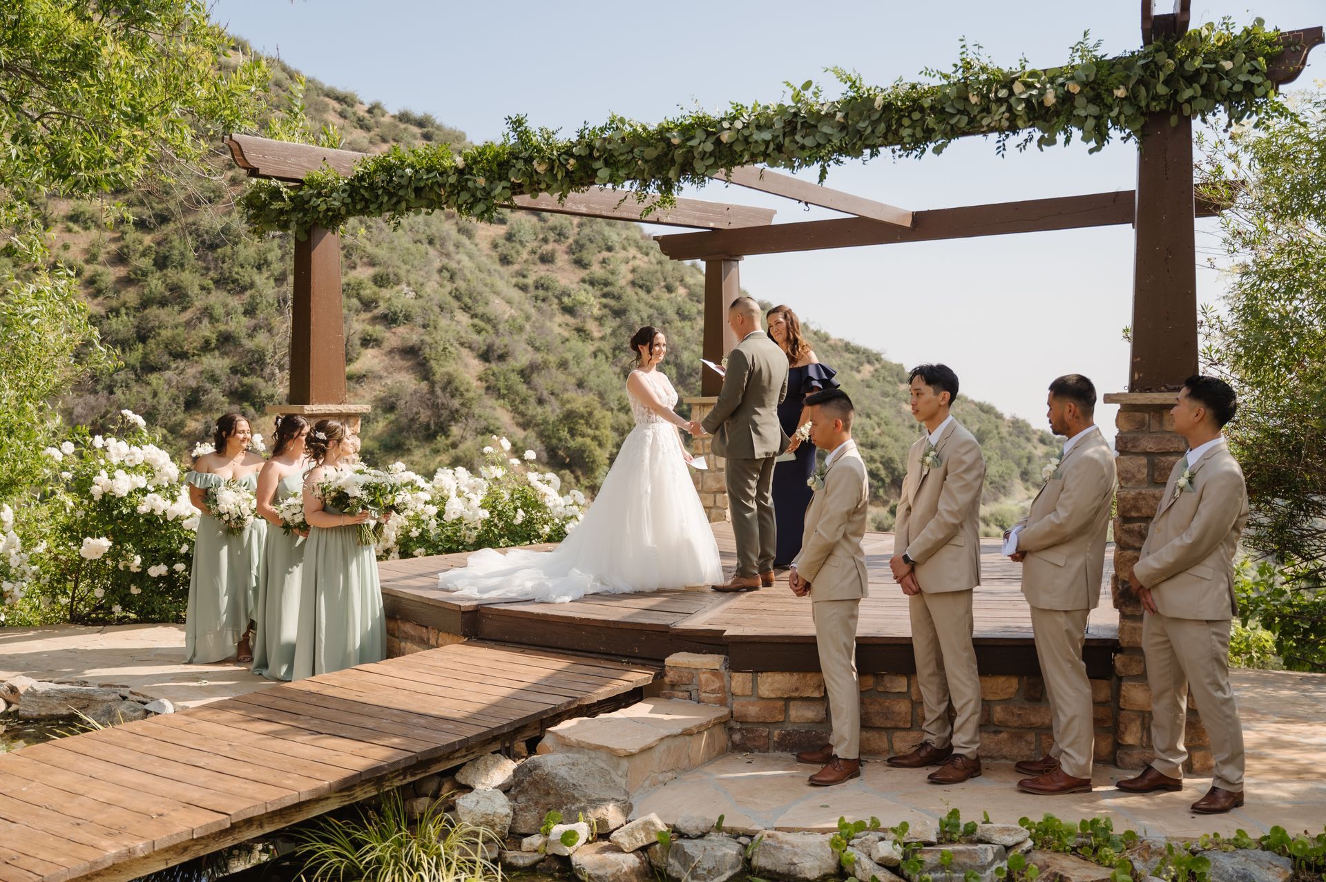 Serendipity wedding ceremony under a vine-covered pergola with bride, groom, bridesmaids, and groomsmen on a stone patio
