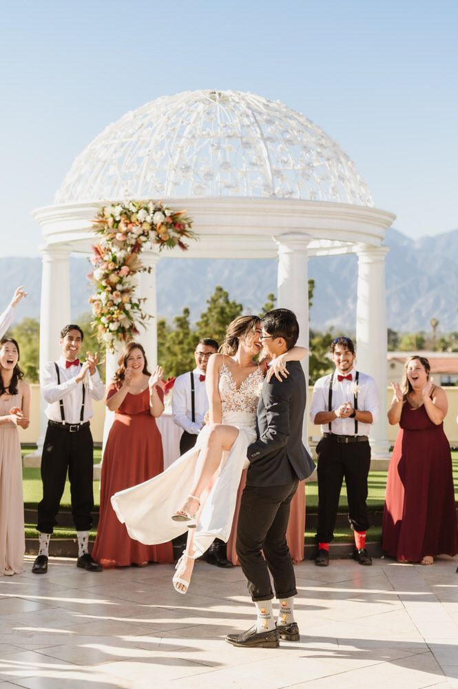 Wedding couple dancing under a white gazebo, with guests clapping in a sunlit outdoor garden.