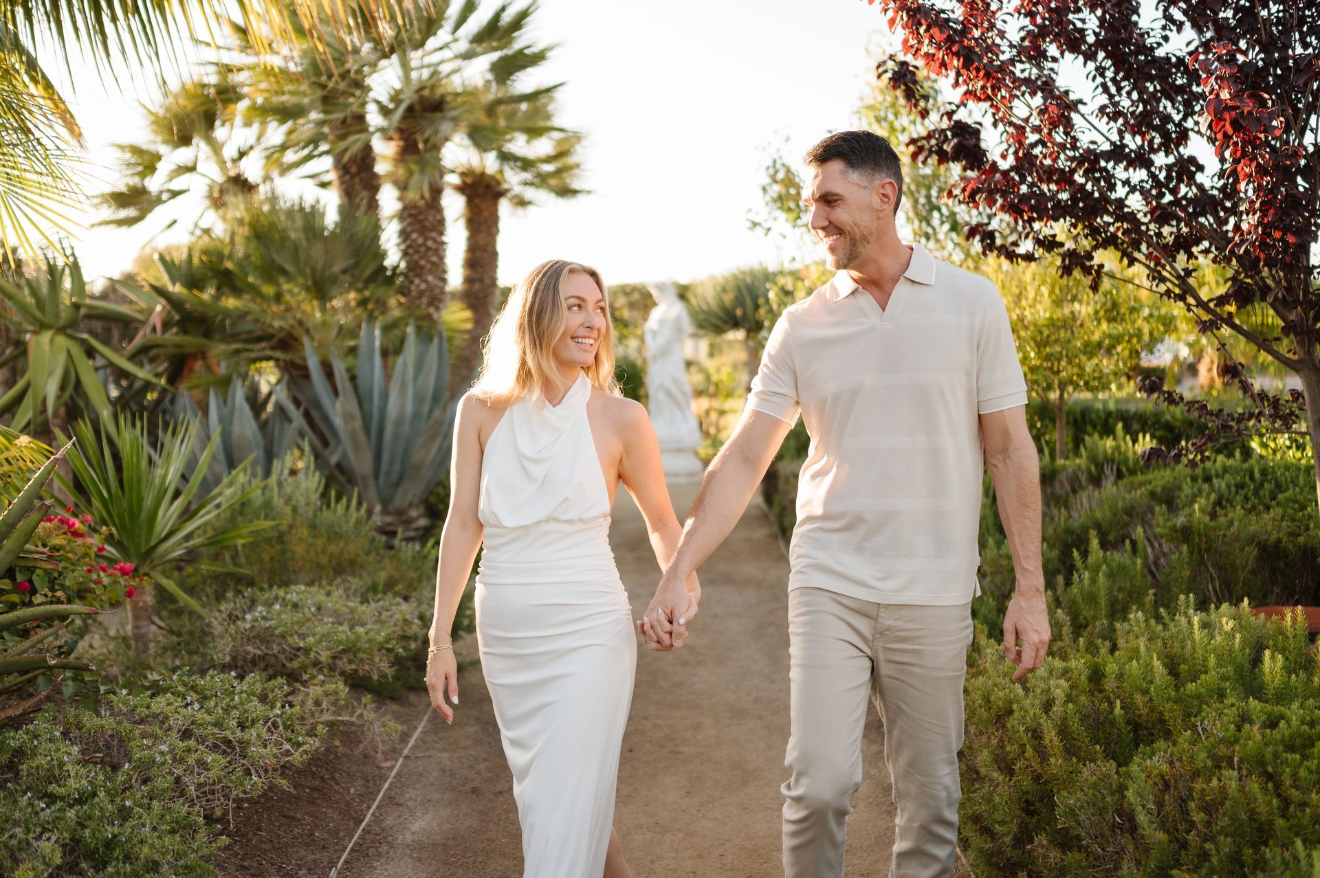 Couple holding hands walking on a garden path with tropical plants at sunset for a proposal with The Yes Girls