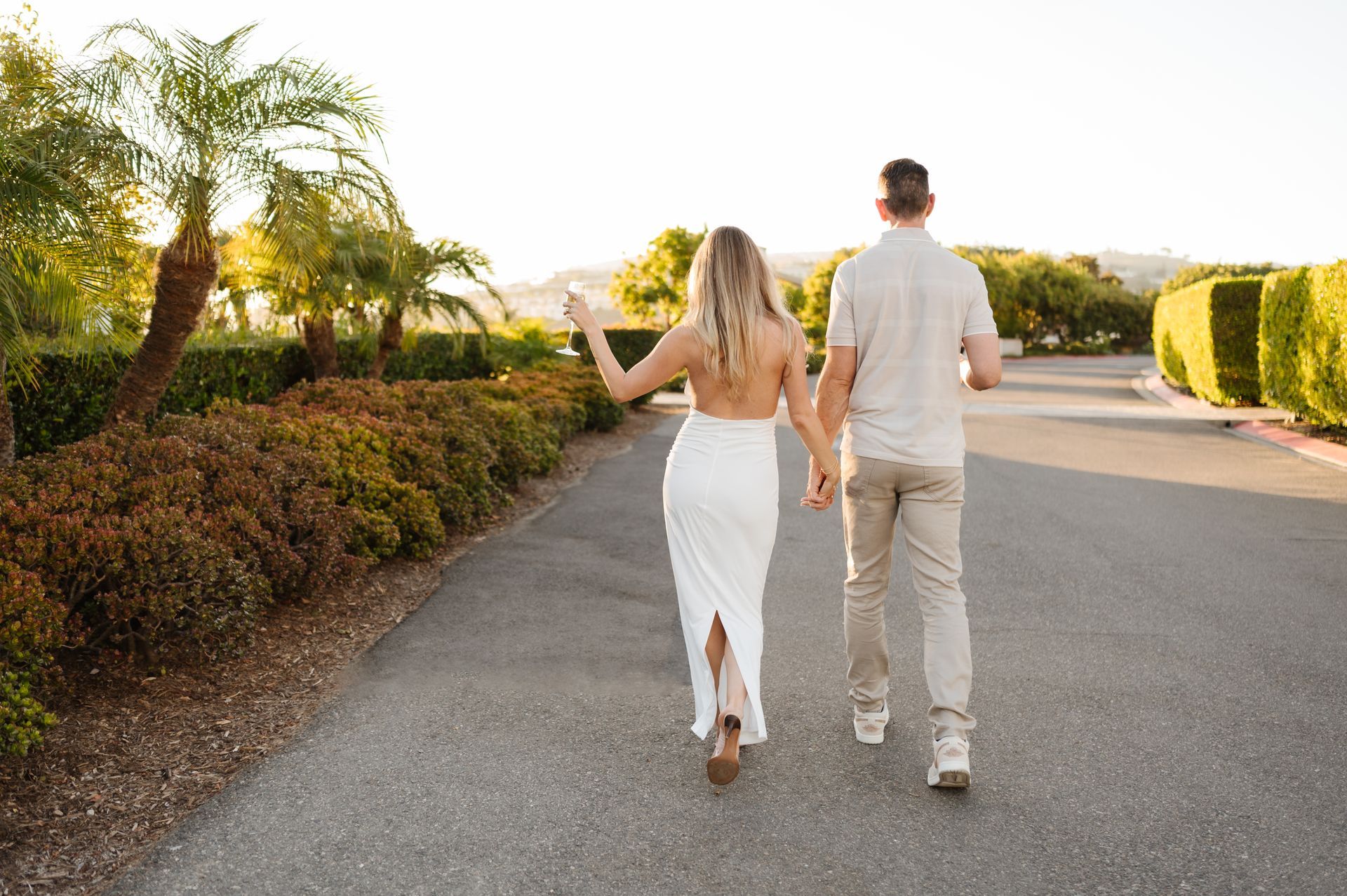 Couple walking hand in hand down a sunny suburban road for a proposal with The Yes Girls, surrounded by palm trees and hedges