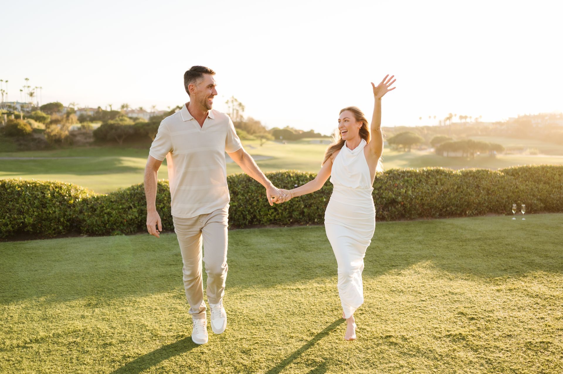 Couple holding hands and smiling while walking on a sunny golf course lawn for a proposal with The Yes Girls