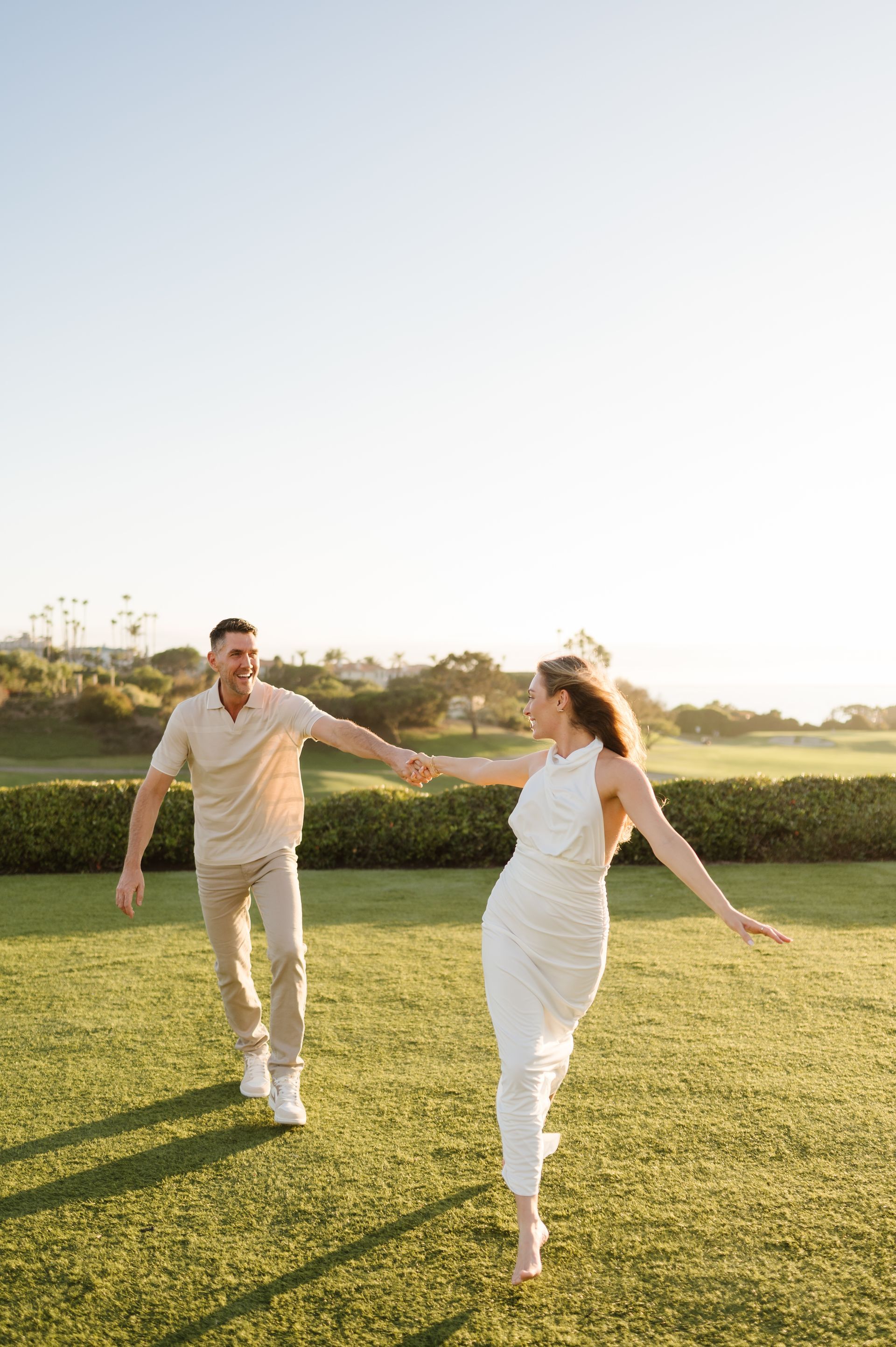 Two people in white outfits dancing hand-in-hand on a sunny lawn outdoors after proposal