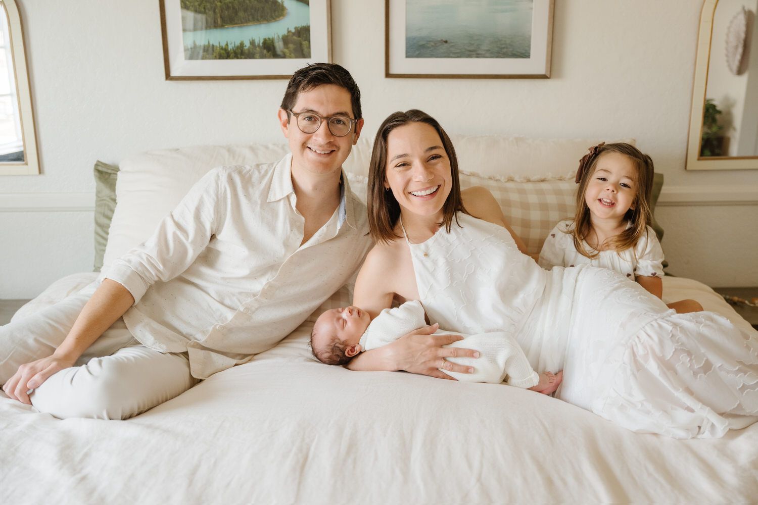 Family of four sitting on a white bed, smiling in a bright bedroom with framed wall art.
