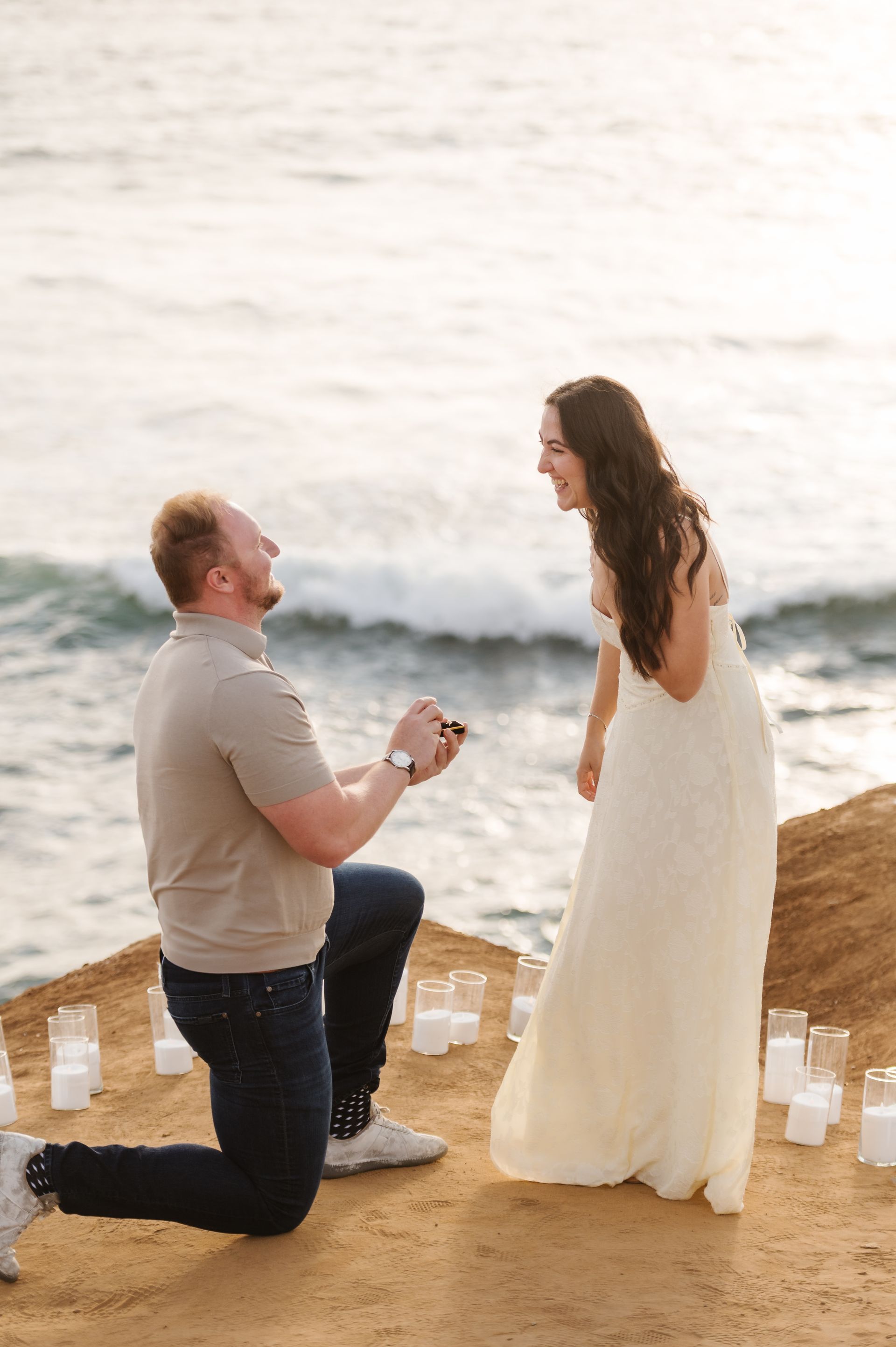 A man kneeling and holding a ring toward a woman in a white dress at sunset for a proposal with The Yes Girls