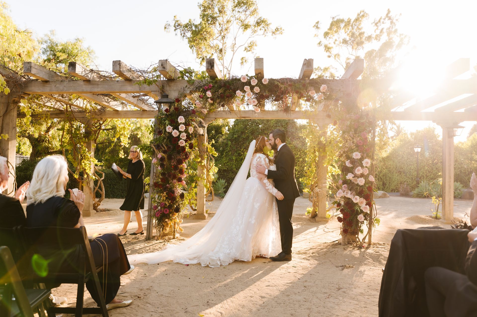 Bride and groom kiss under a flower-covered pergola at an outdoor sunset wedding ceremony