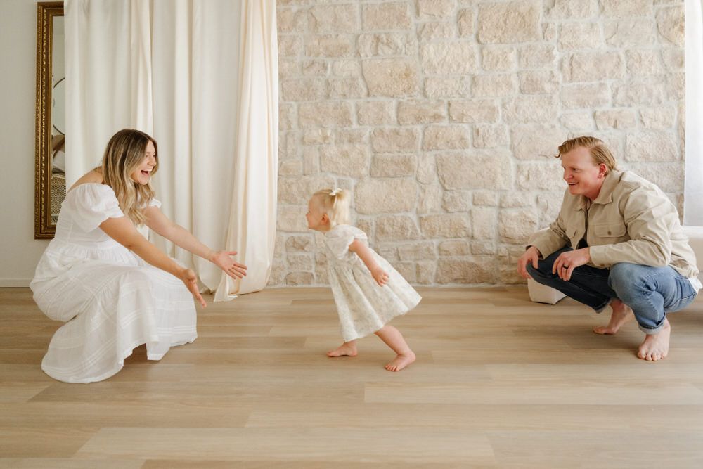 Woman and toddler crouch on a wooden floor beside a stone wall, facing a small child in a white dress.