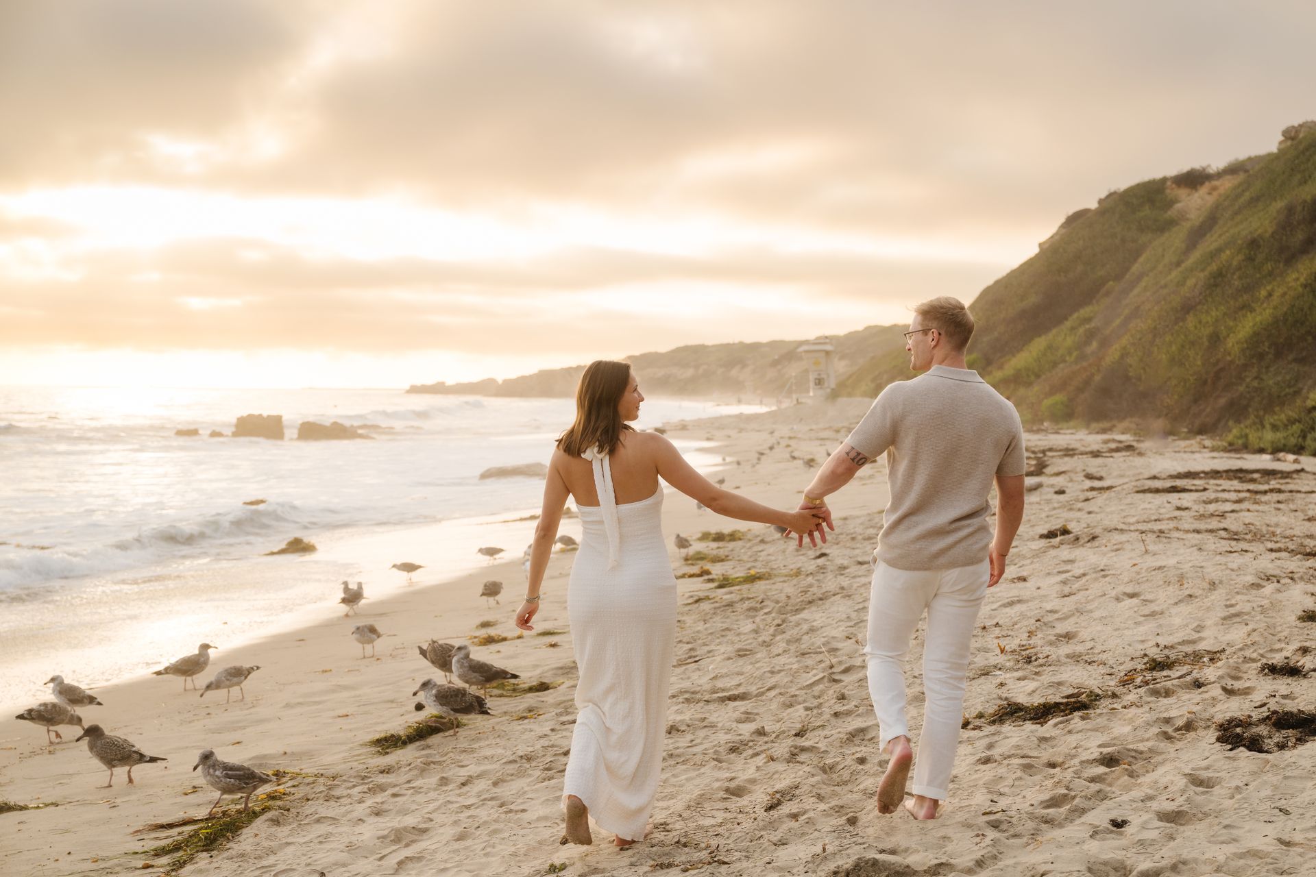 Couple holding hands while walking along a sandy beach at sunset for a proposal with The Yes Girls