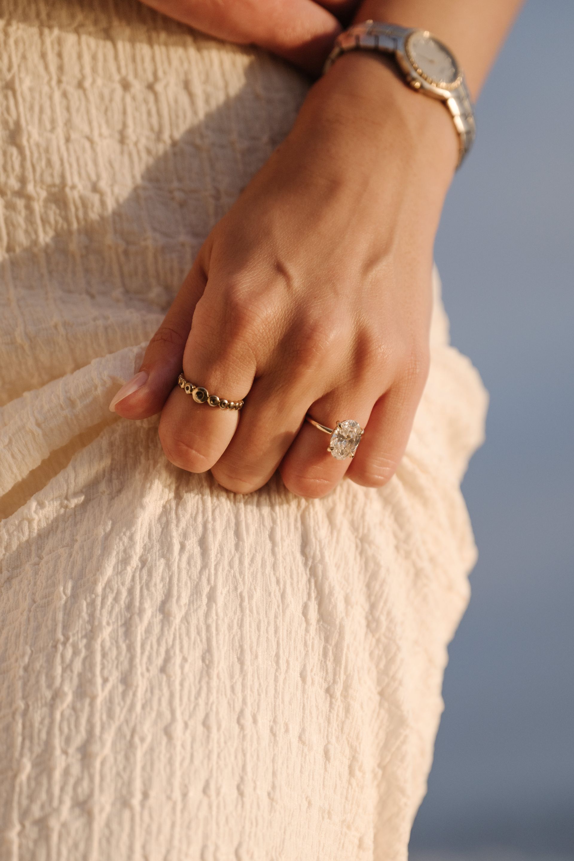 Hand resting on a cream skirt, wearing rings and a wristwatch against a soft blue background