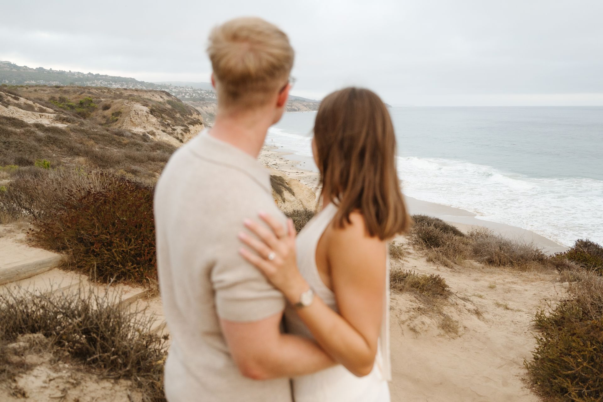 Couple standing on a coastal cliff, looking out over the ocean on an overcast day for a proposal with The Yes Girls