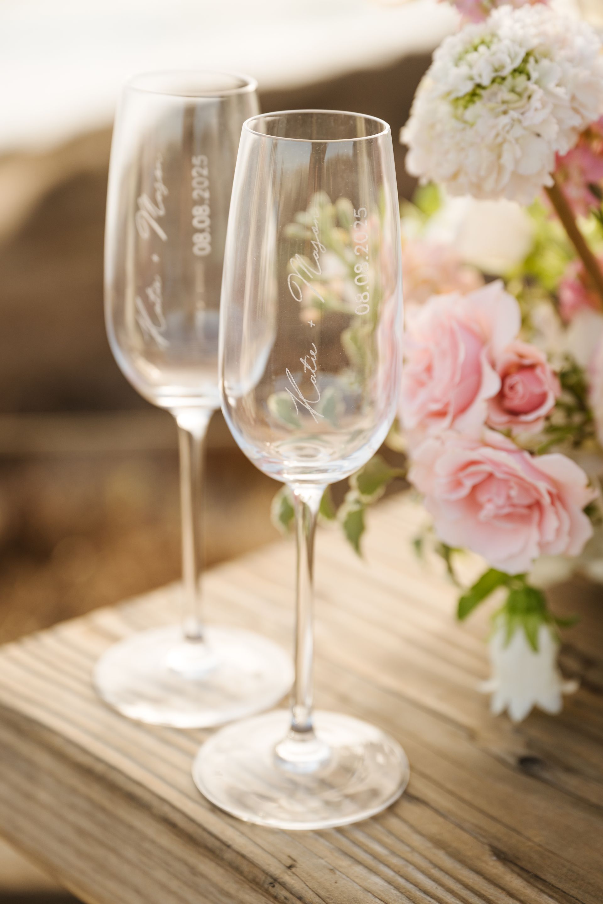Two champagne flutes on a wooden table beside pink and white flowers for proposal with The Yes Girls