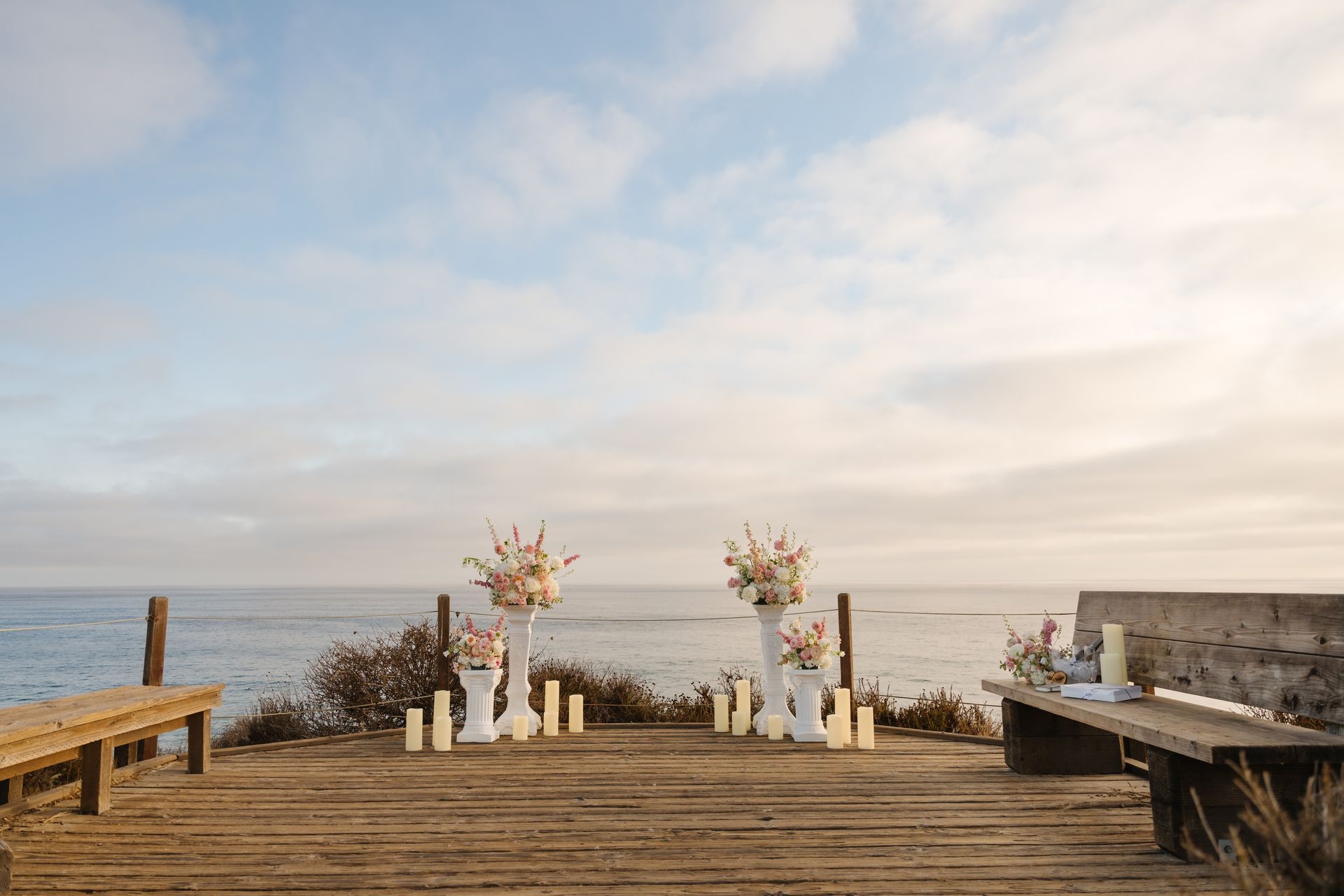 Decorated wooden deck overlooking the ocean with white floral arrangements and candles at sunset