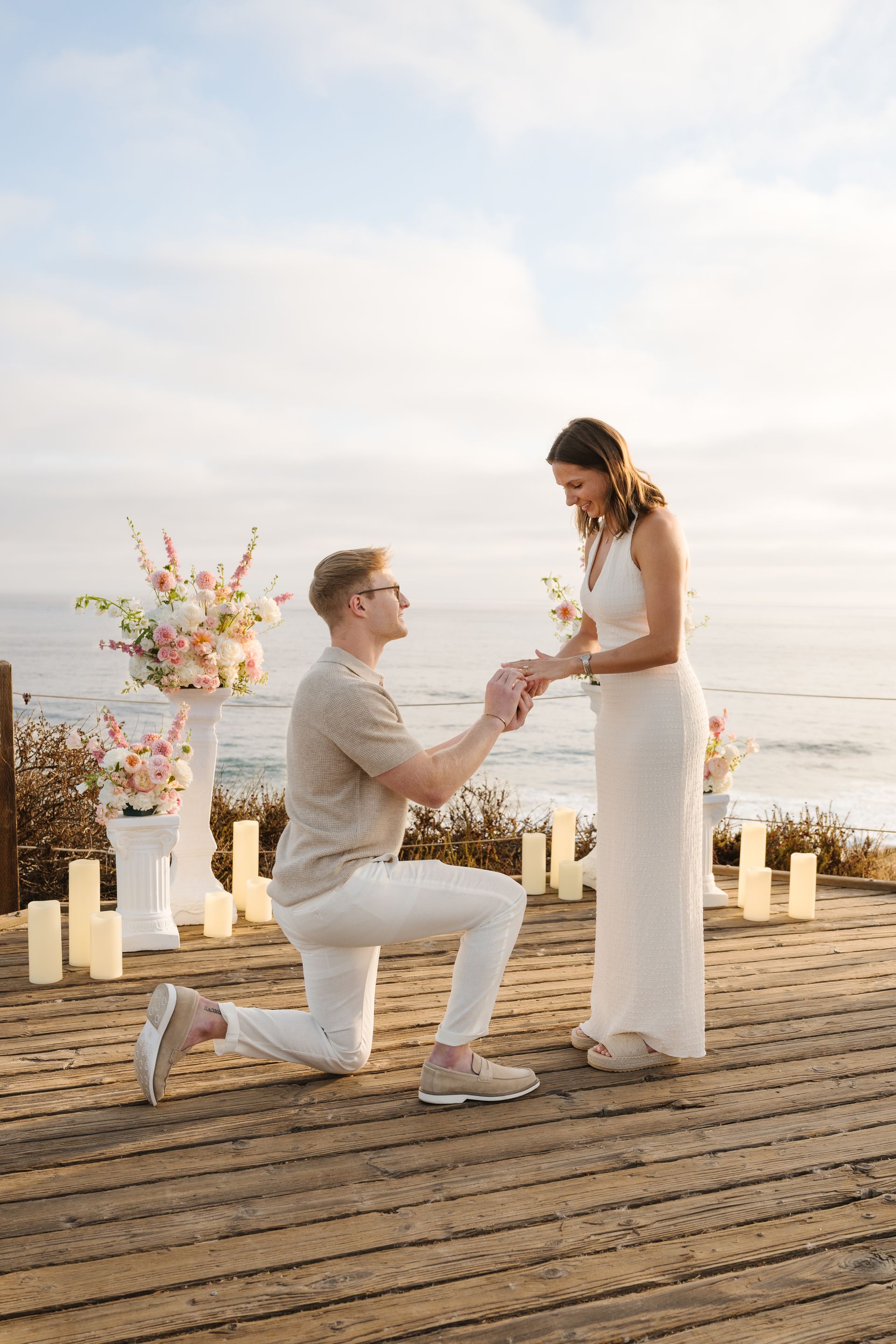 Man kneels to propose to a woman in white on a beach at sunset with The Yes Girls