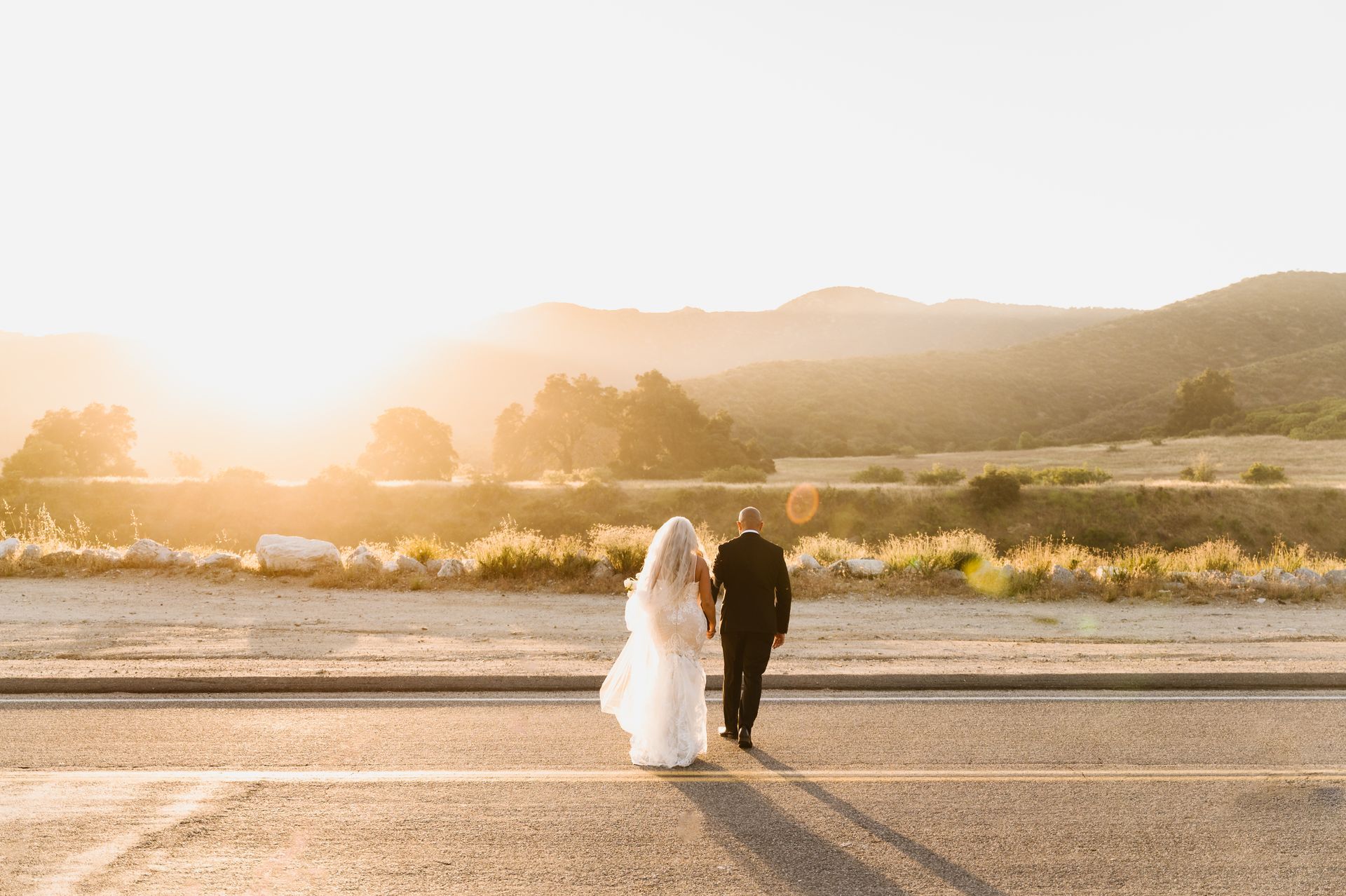 Bride and groom walking hand in hand on a road at sunset with mountains in the distance