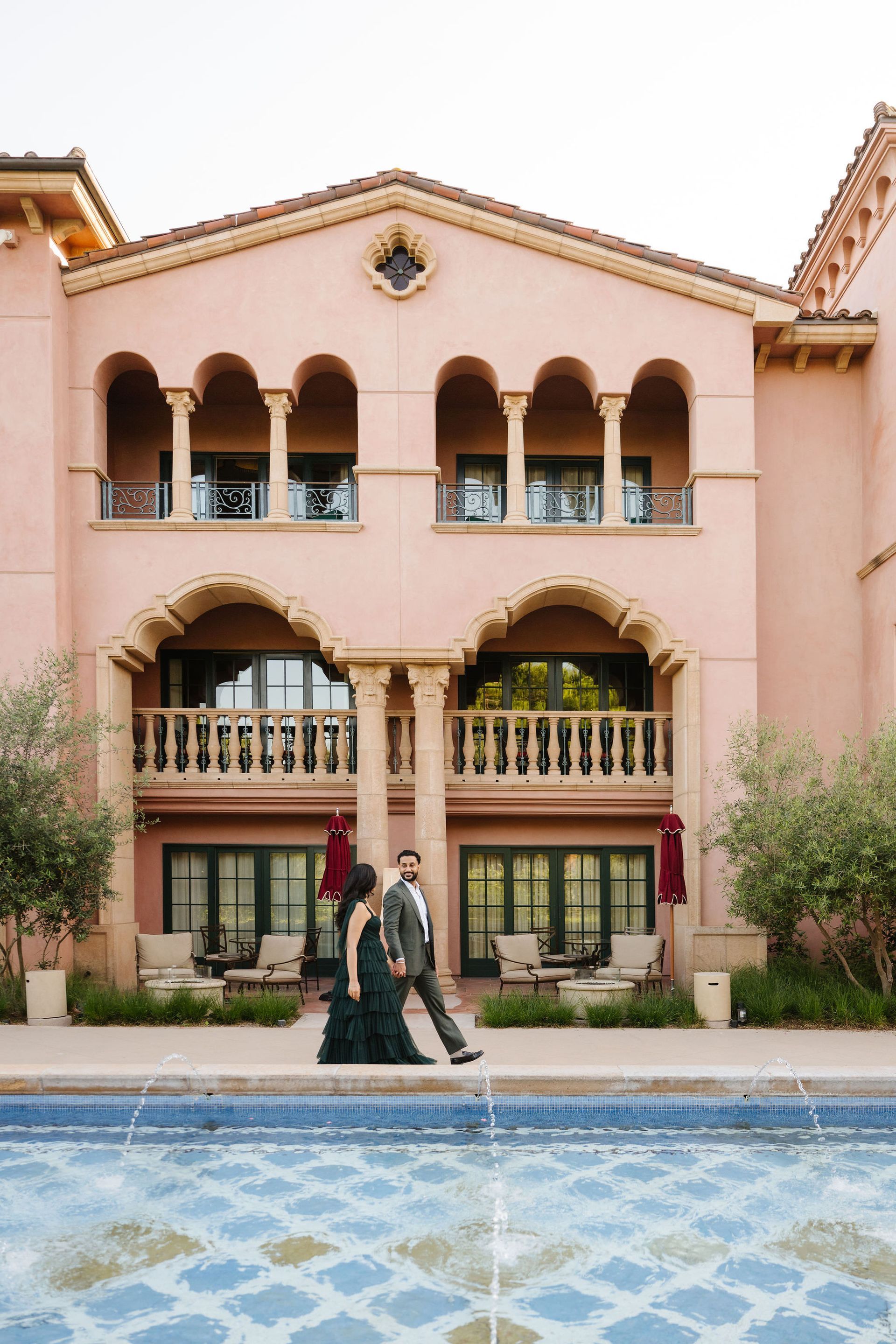 Two people walking beside a blue pool in front of a pink Spanish-style building for a proposal with The Yes Girls