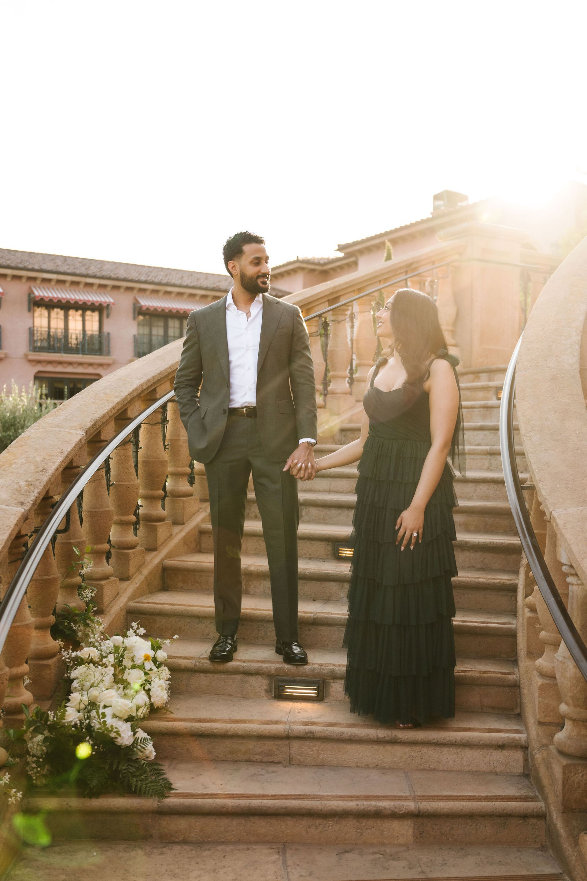 Couple holding hands on stone steps, with floral decorations and warm sunlight at an outdoor venue.