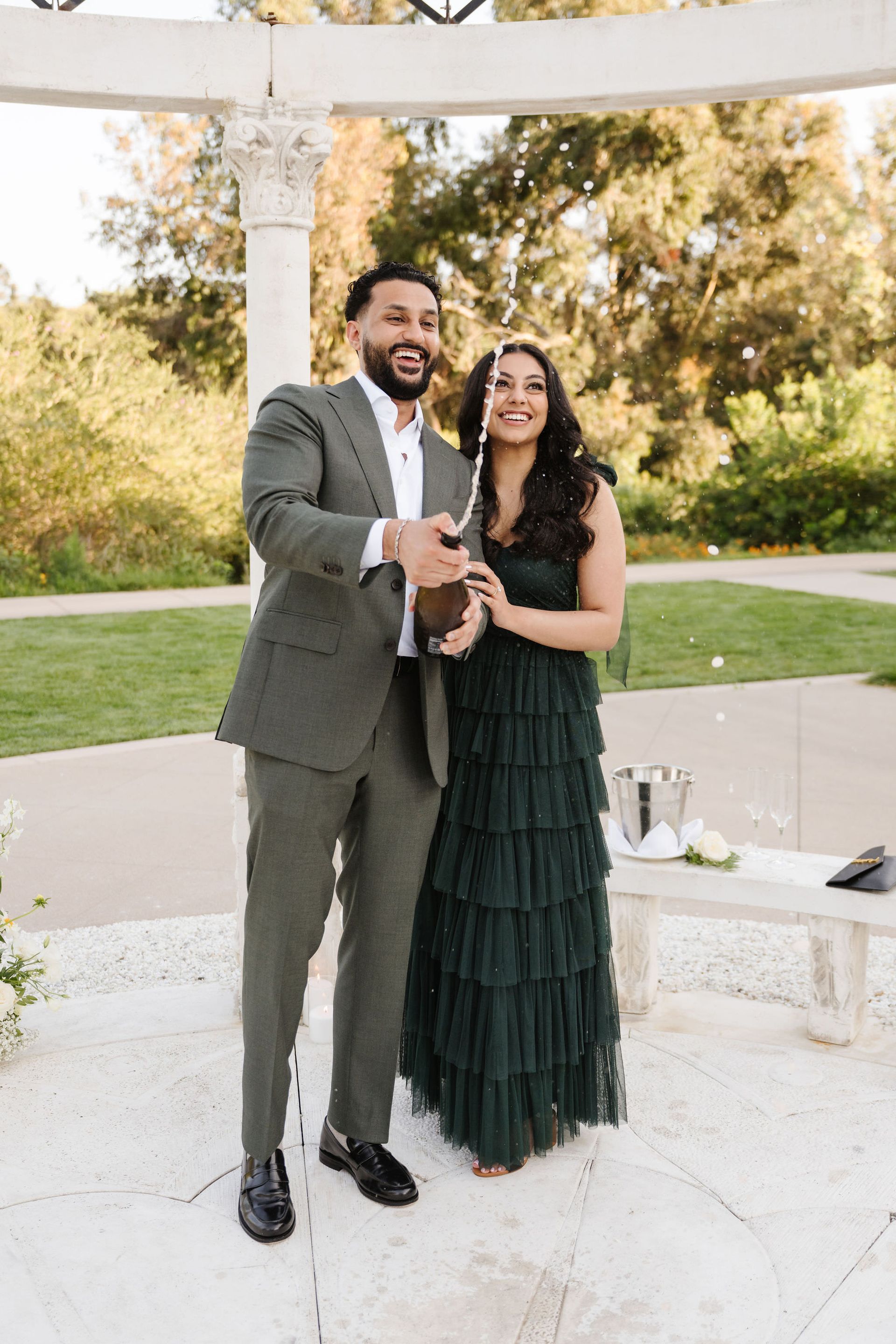 Couple in formal attire standing outdoors under a white gazebo for a proposal with The Yes Girls, smiling and holding hands.