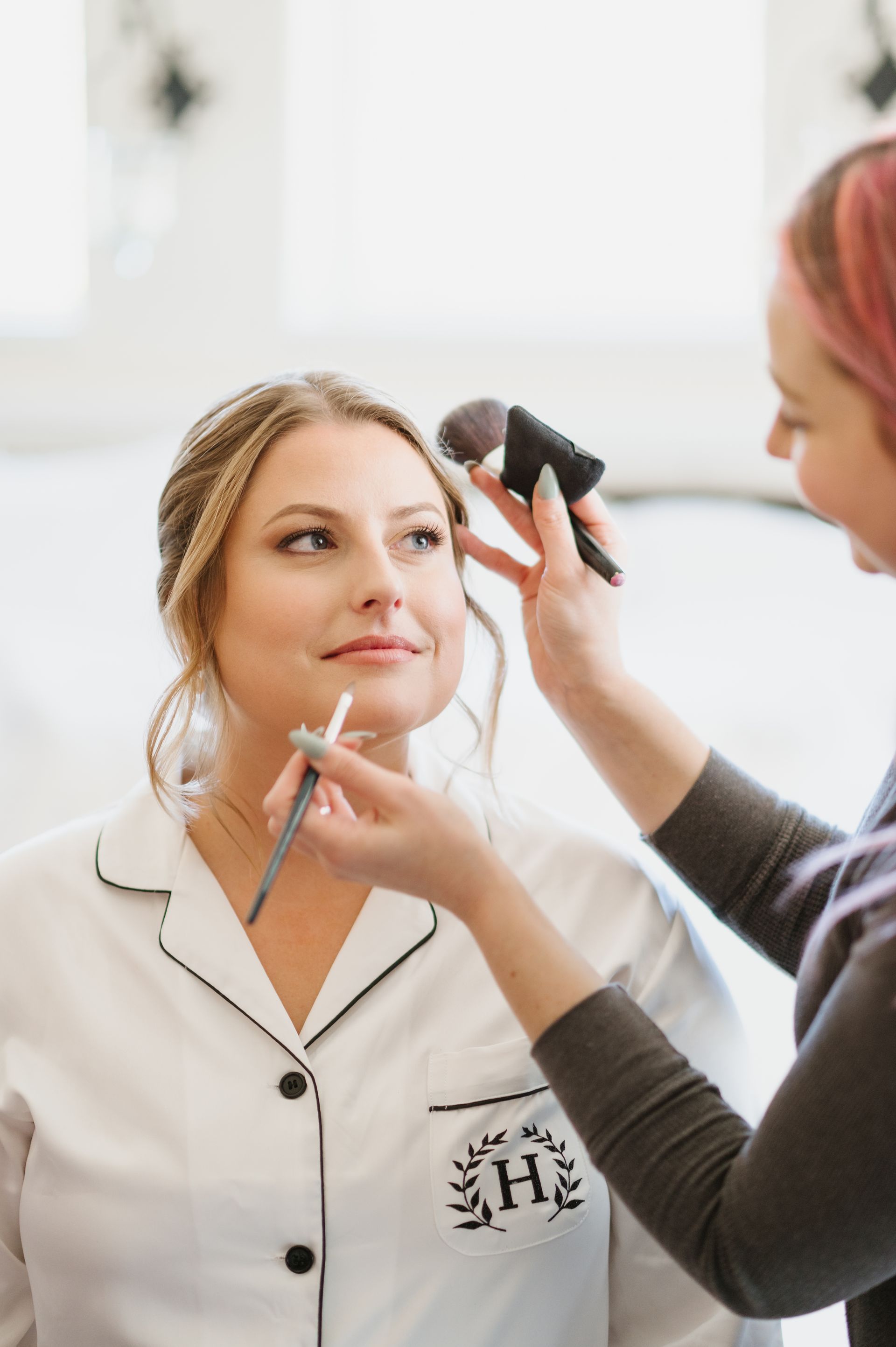 A makeup artist applying makeup to a client wearing white monogrammed pajamas.