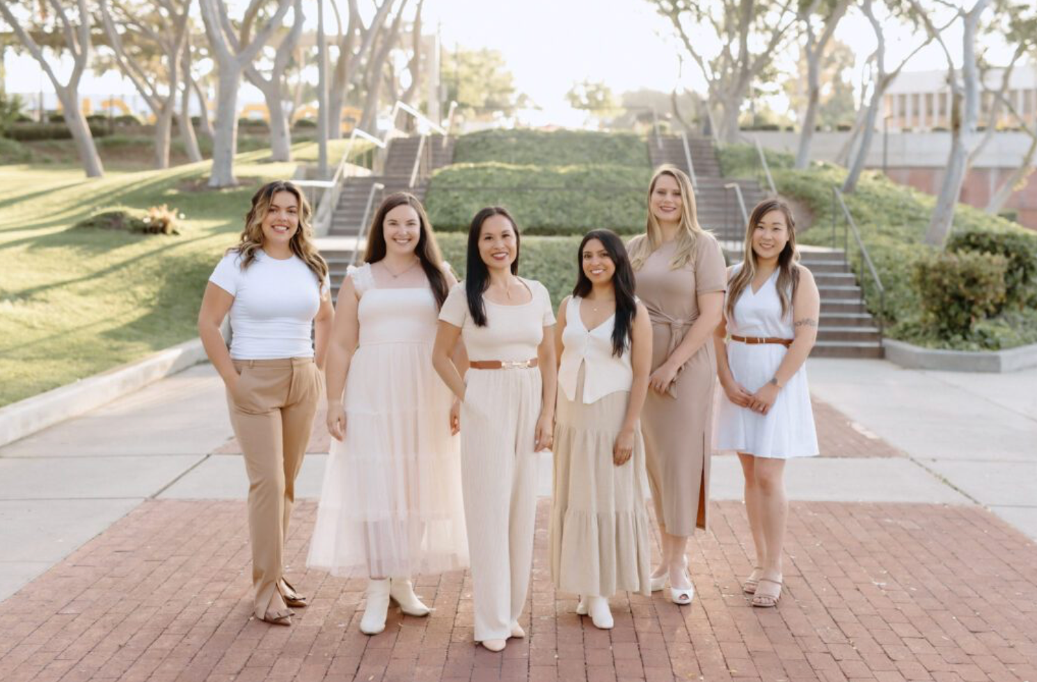 Six wedding planners in light-colored clothing stand together on a brick walkway outdoors