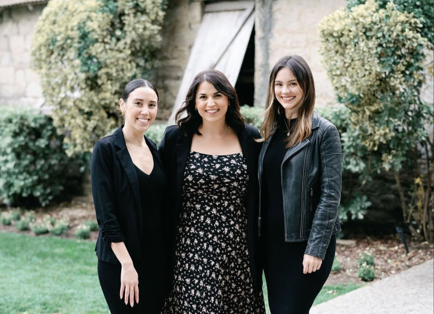 Three wedding planners stand side-by-side on a stone path in front of a building and greenery