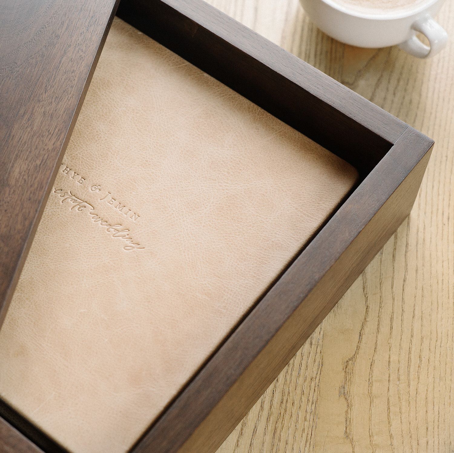 Top-down view of a wedding album in a wood box on a light wood table beside a white cup