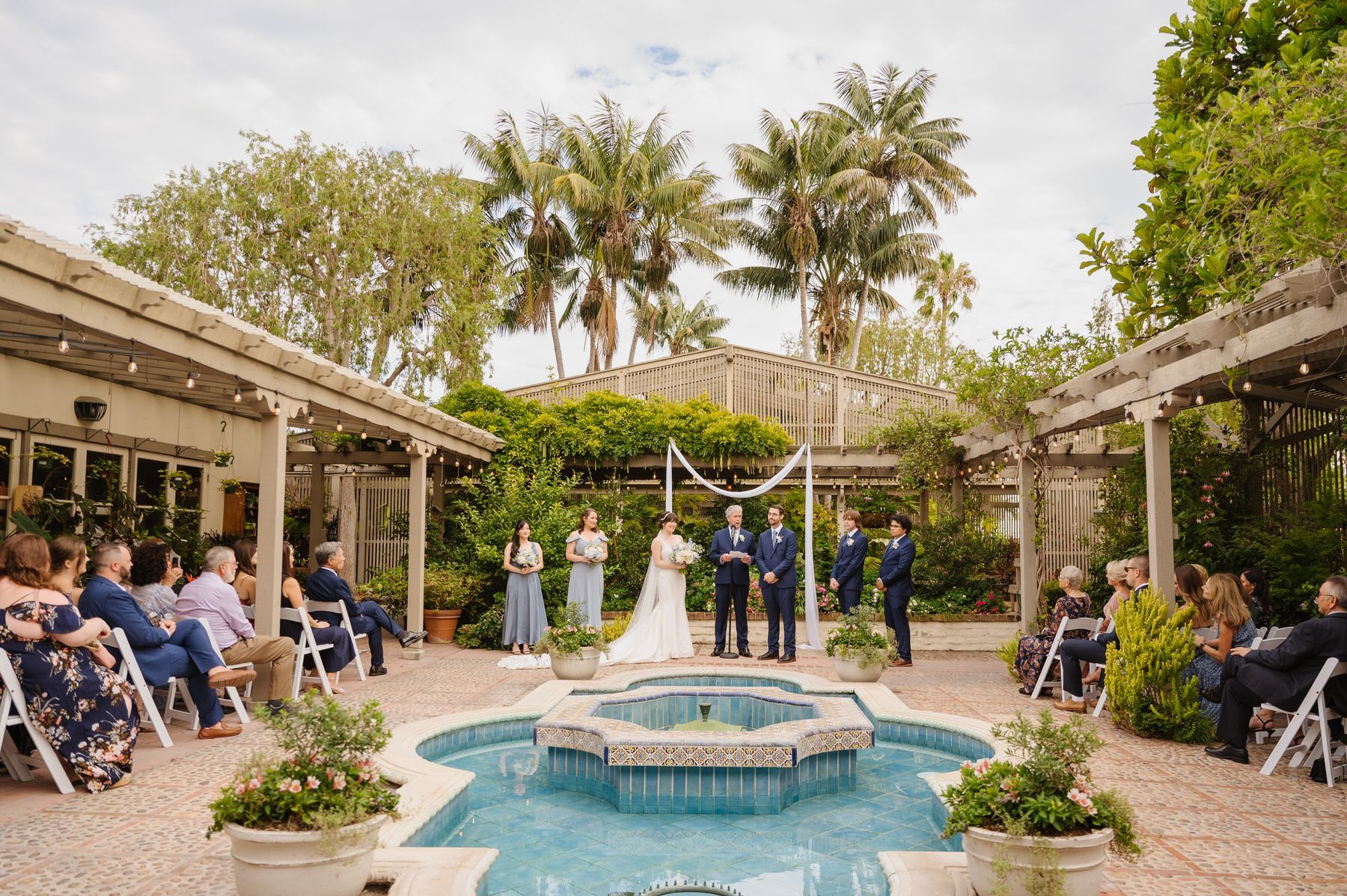 A bride and groom are standing in front of a fountain during their wedding ceremony.