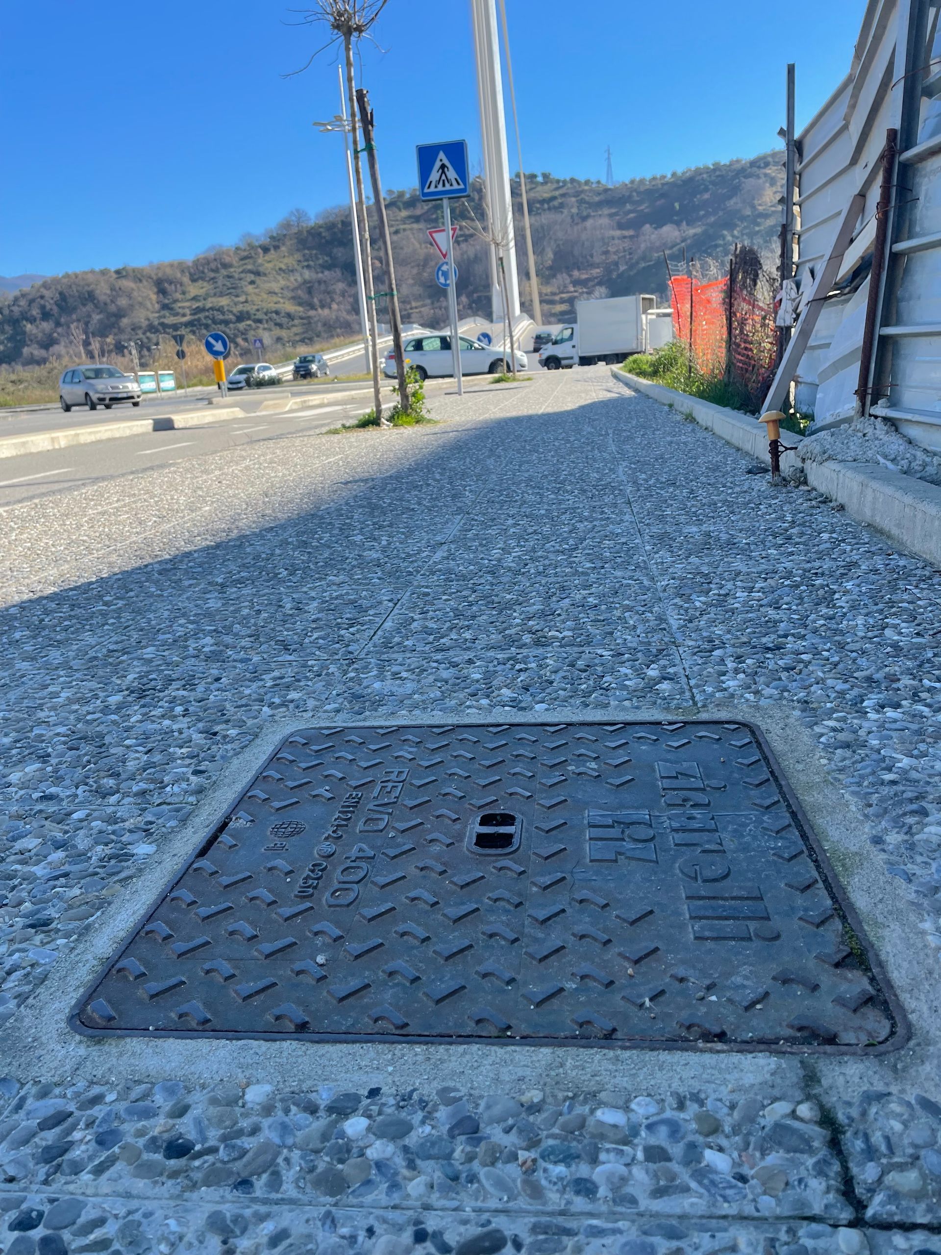 Marciapiede in ghiaia con un tombino accanto a una strada, con le montagne sullo sfondo.