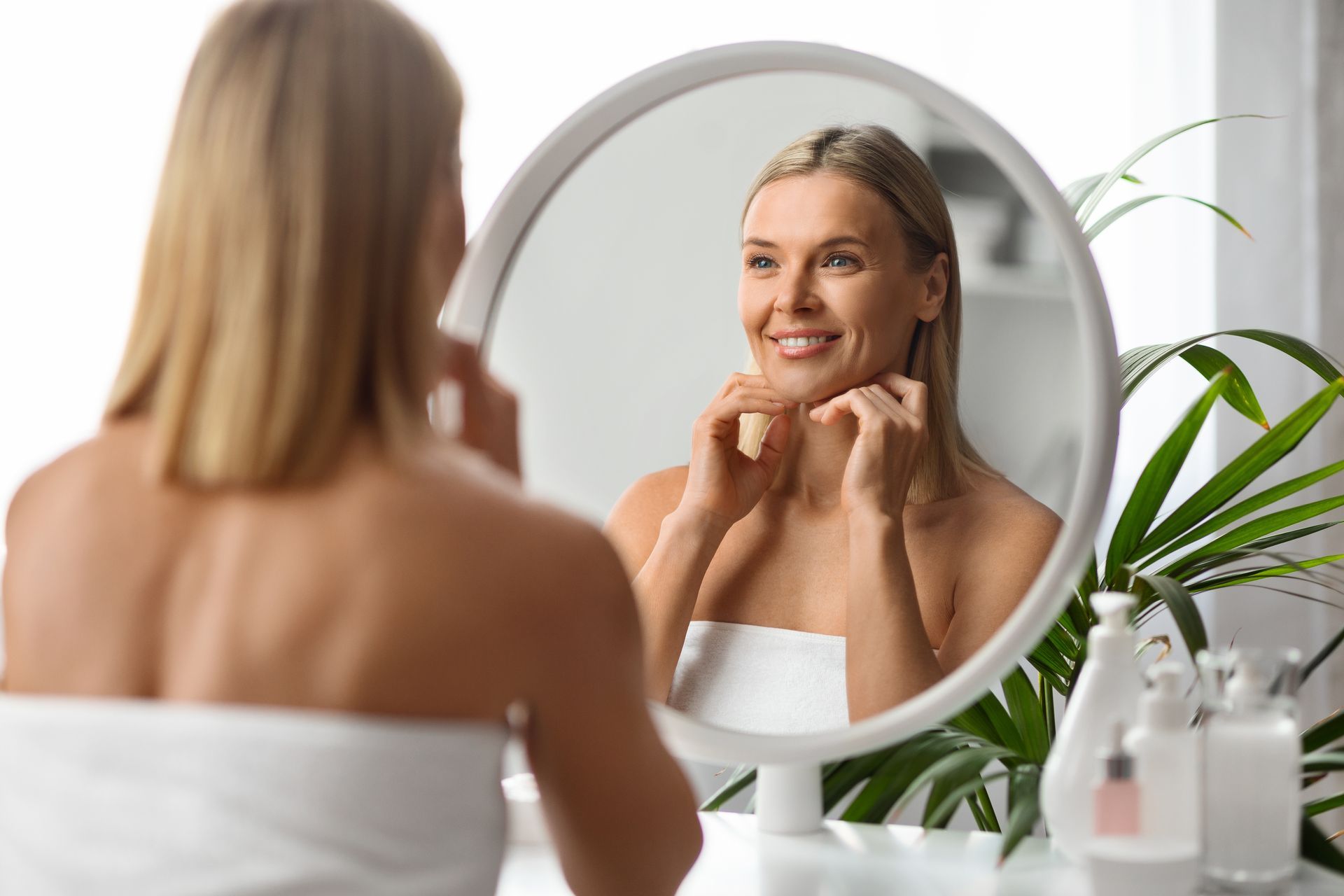 Blonde woman in towel looking in mirror, touching her chin and smiling