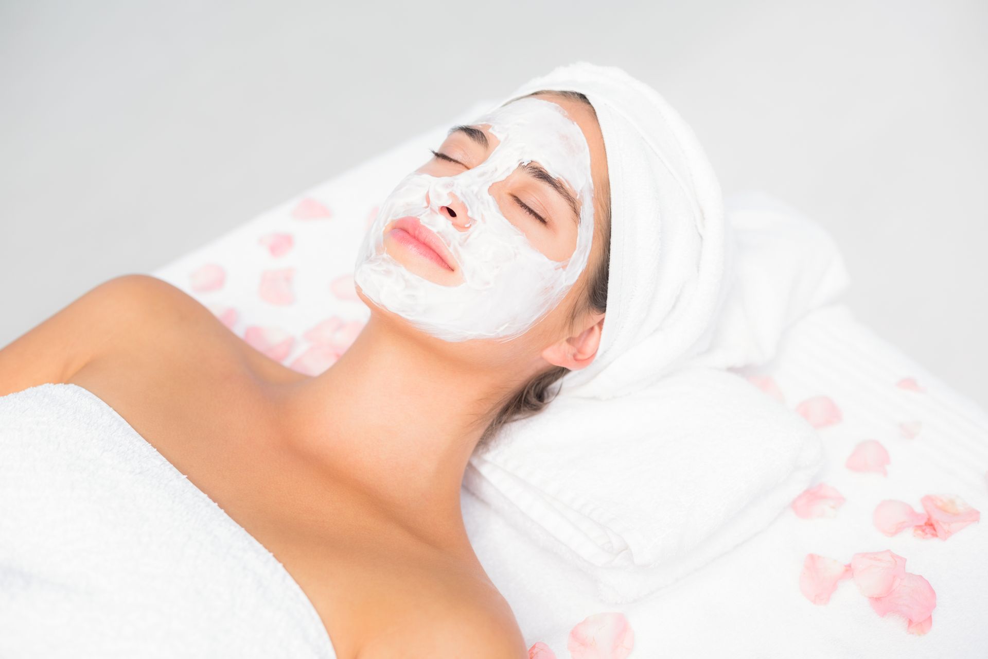 Woman receiving a white facial mask while lying on a spa bed with a towel wrapped around her head