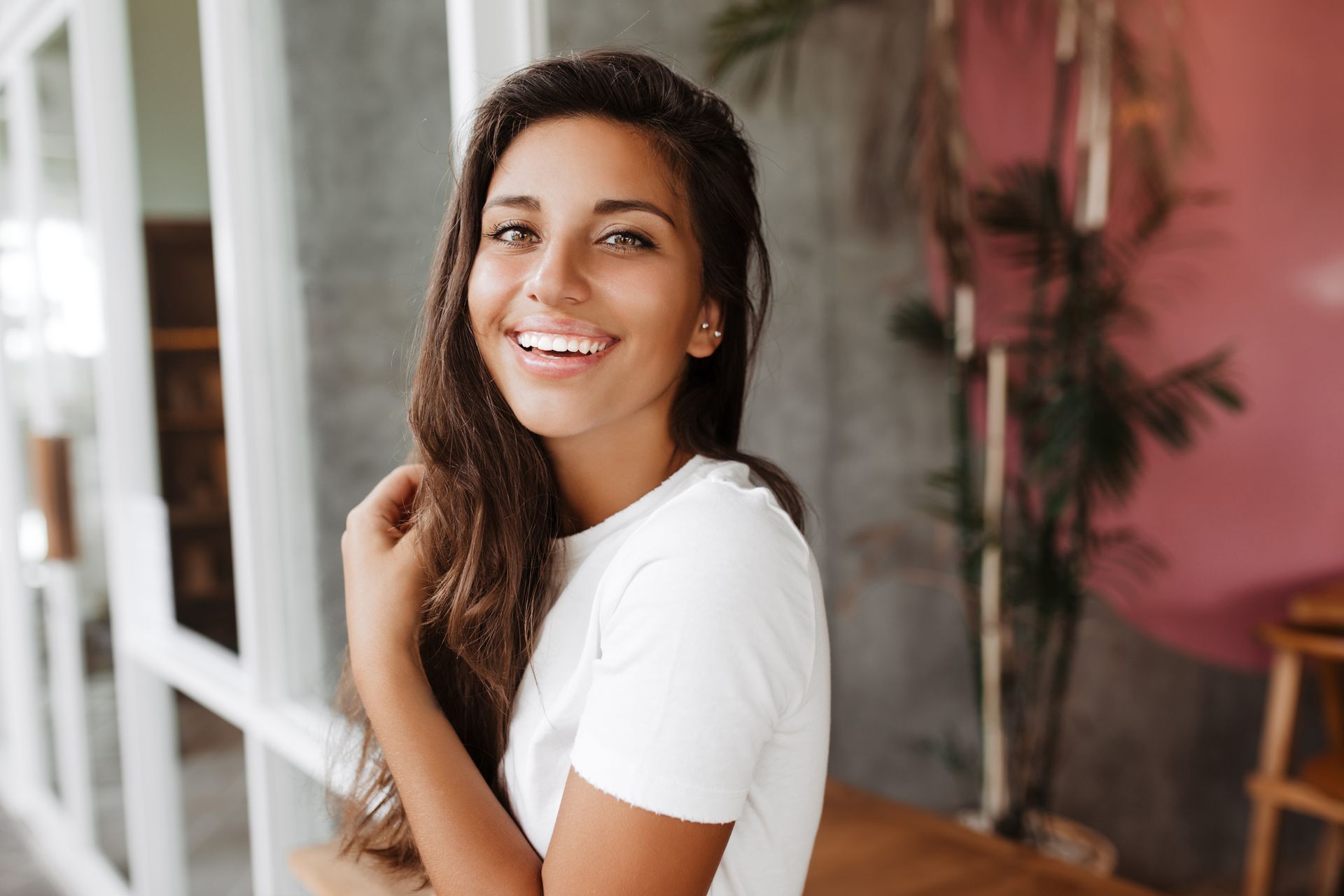 A woman in a white t-shirt is smiling while sitting at a table in front of a window.
