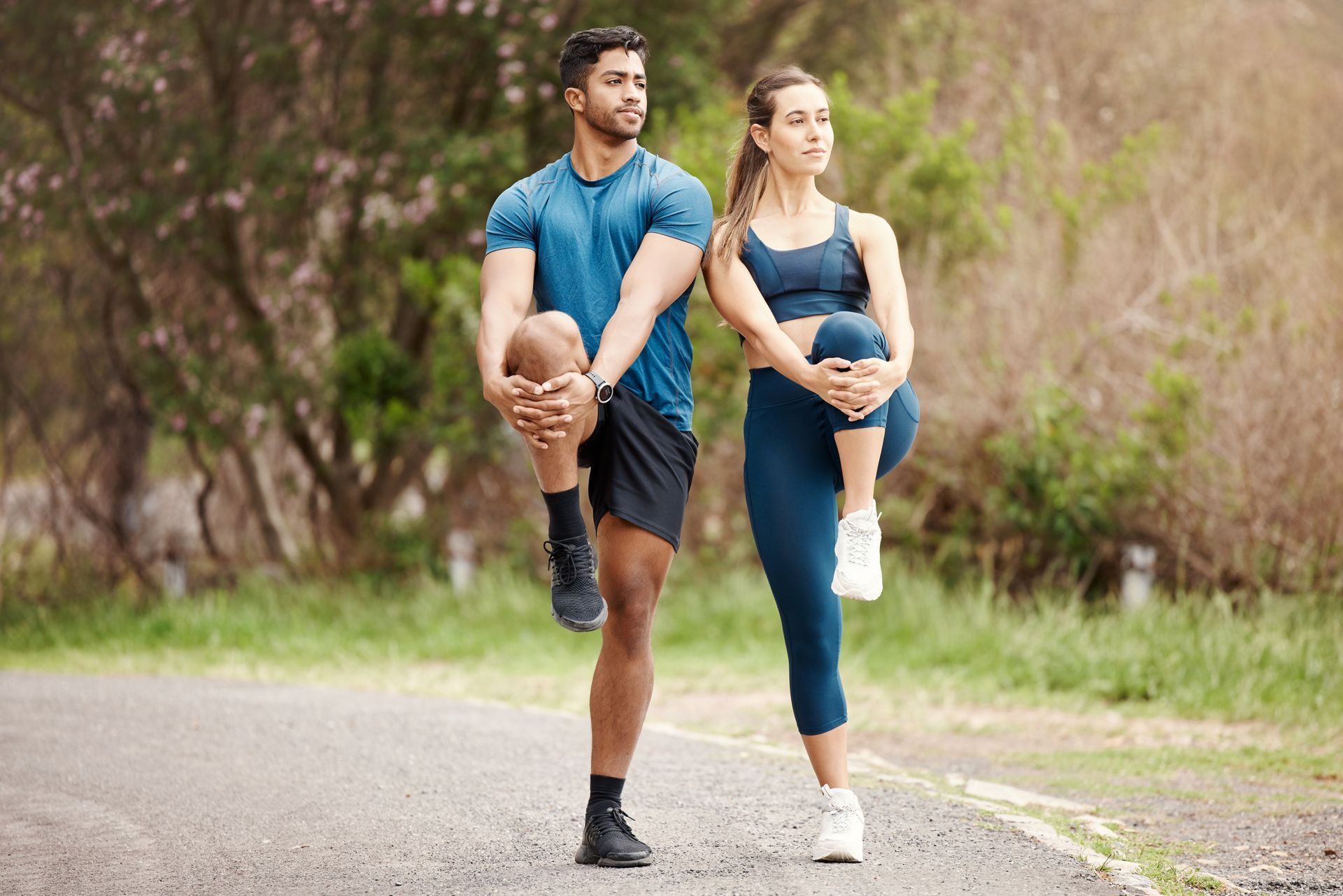 Two runners stretching on a path in a green park, wearing blue athletic clothes.