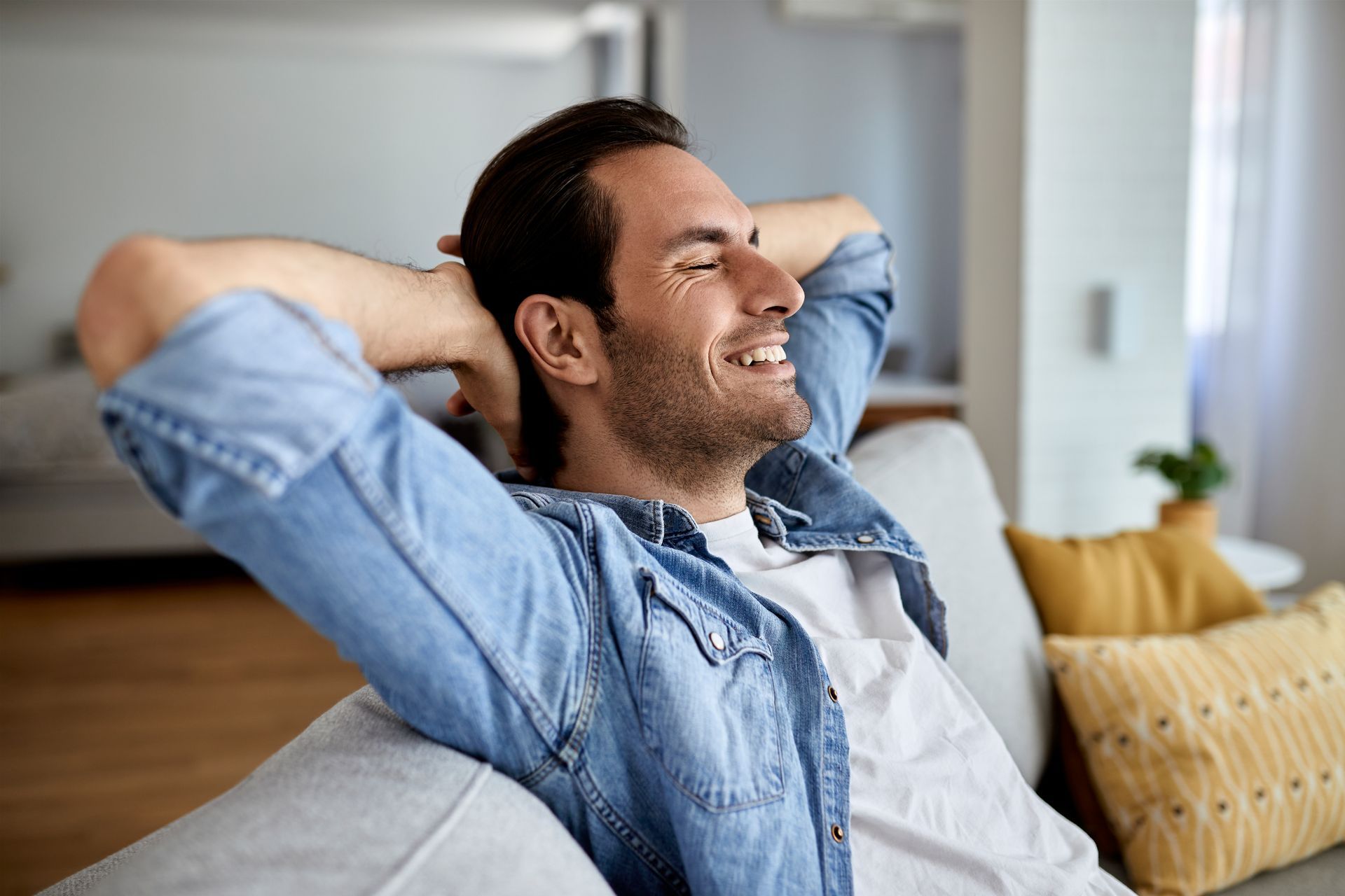 Relaxing man leaning back on a couch with hands behind his head, smiling in a bright living room