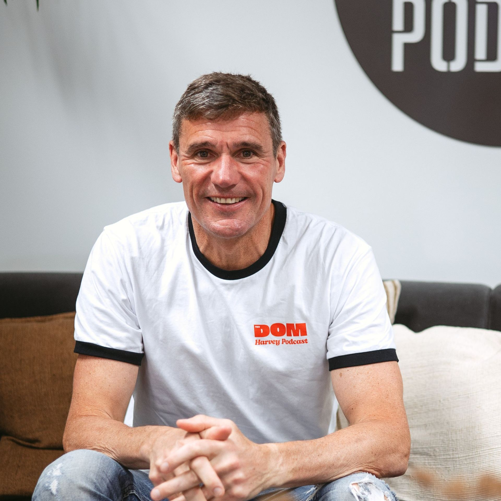 Man in a white T-shirt with black trim sits on a couch, smiling in a modern office lounge.