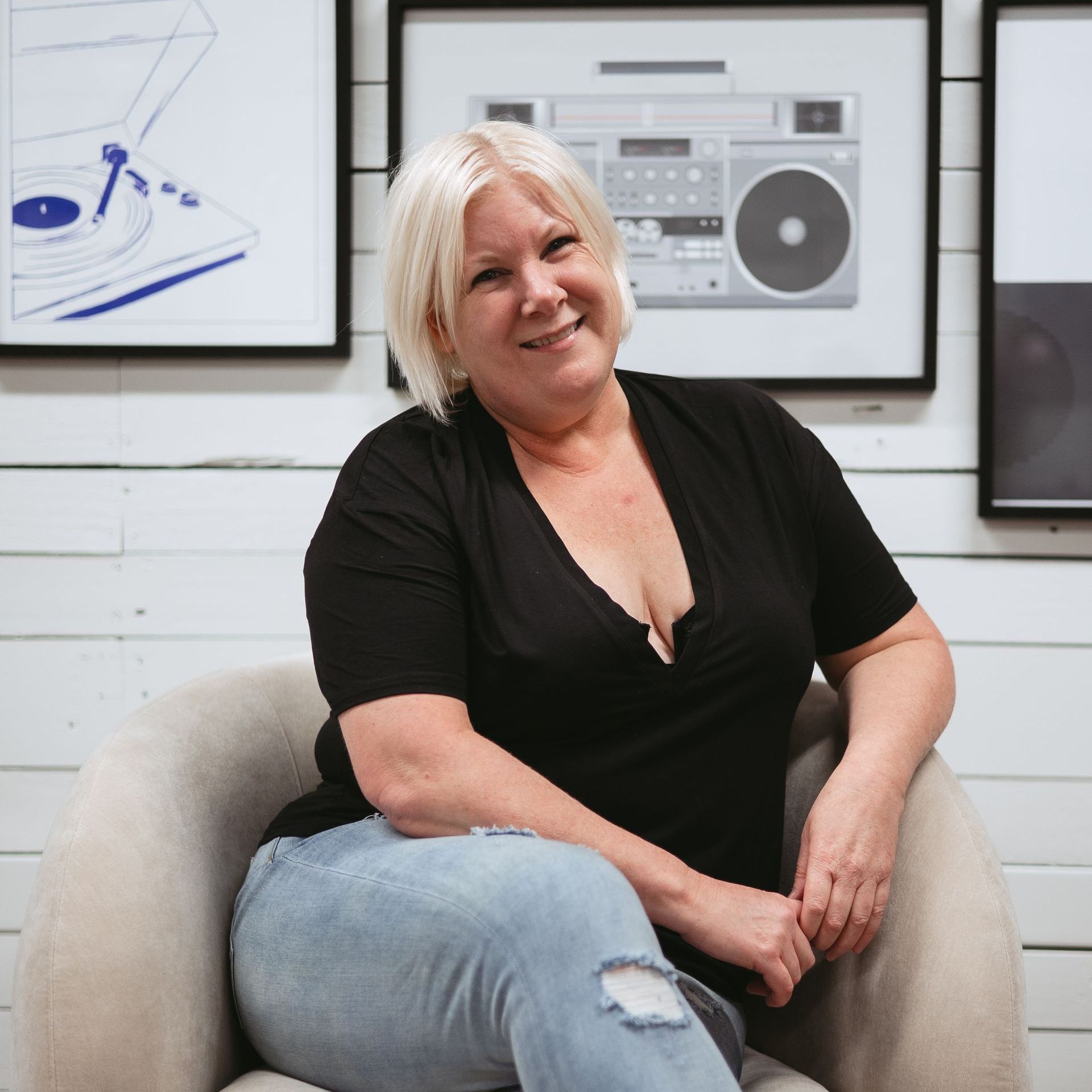 Smiling woman in a black top sitting in a beige chair against framed wall art.