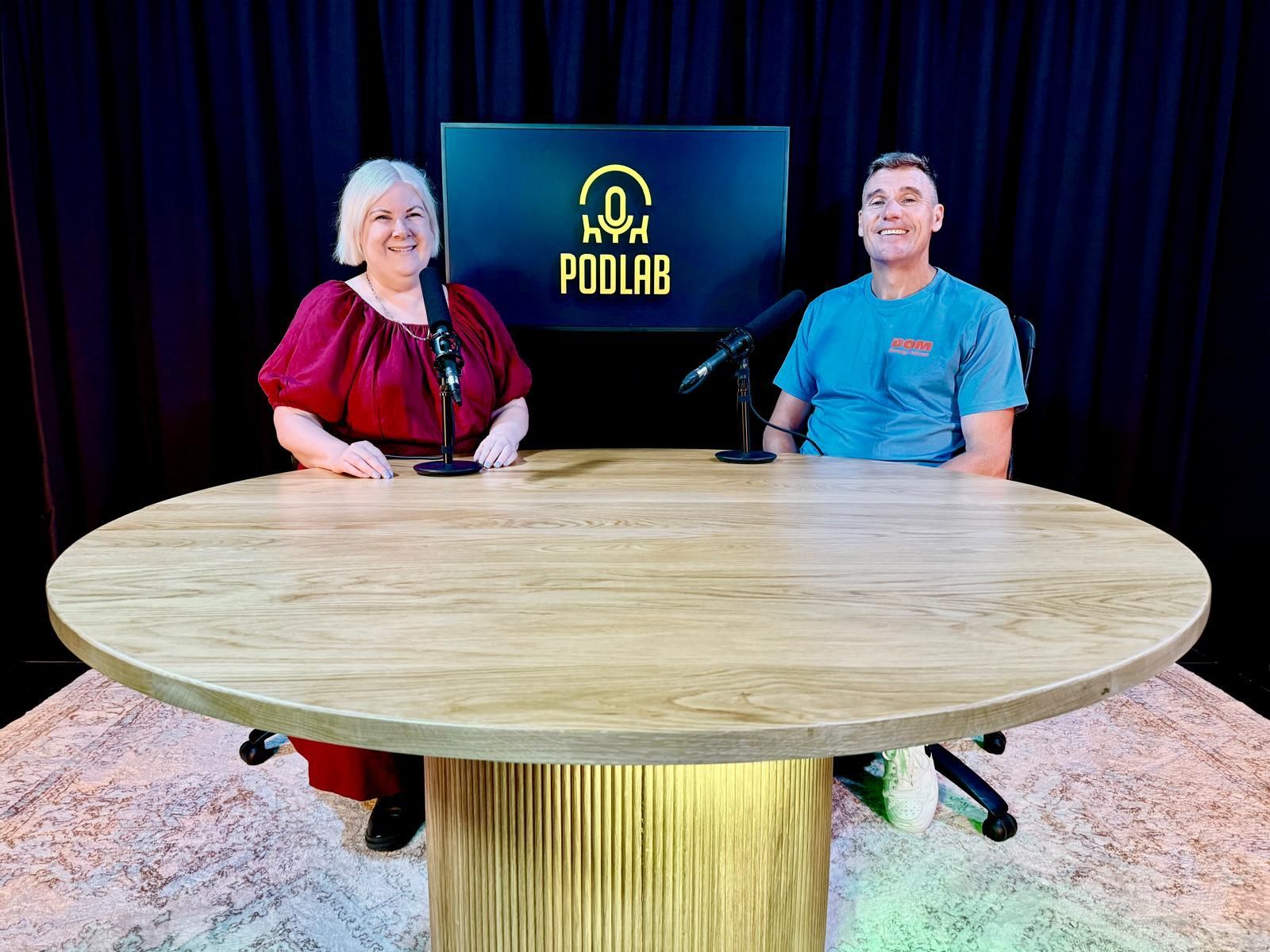Two people seated behind a round wooden table in a studio with a POOLAB sign and dark curtains.