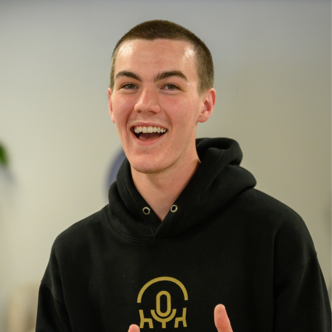 Young person in a black PODLAB hoodie, standing and smiling indoors.