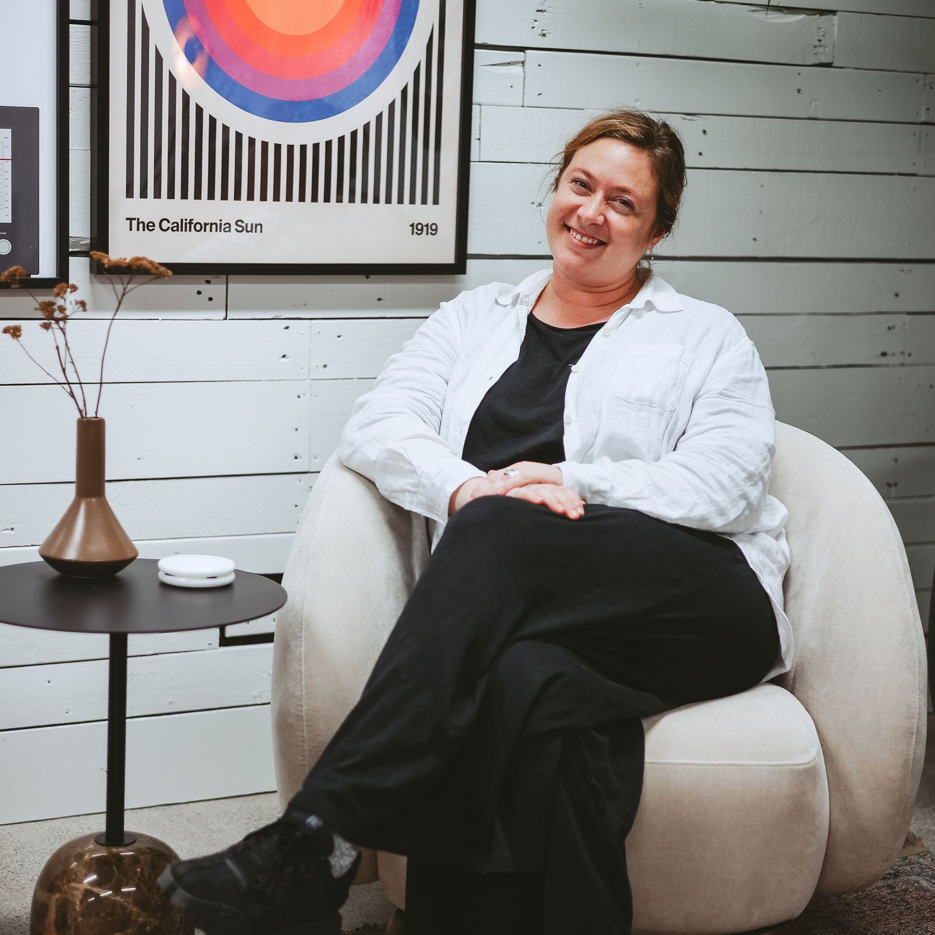 Woman sitting in a beige chair beside a small table and vase, smiling in a modern room with framed wall art.