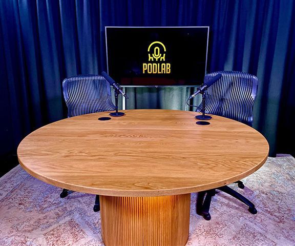 Podcast studio with round wooden table, microphones, chairs, and a screen displaying the PODLAB logo.