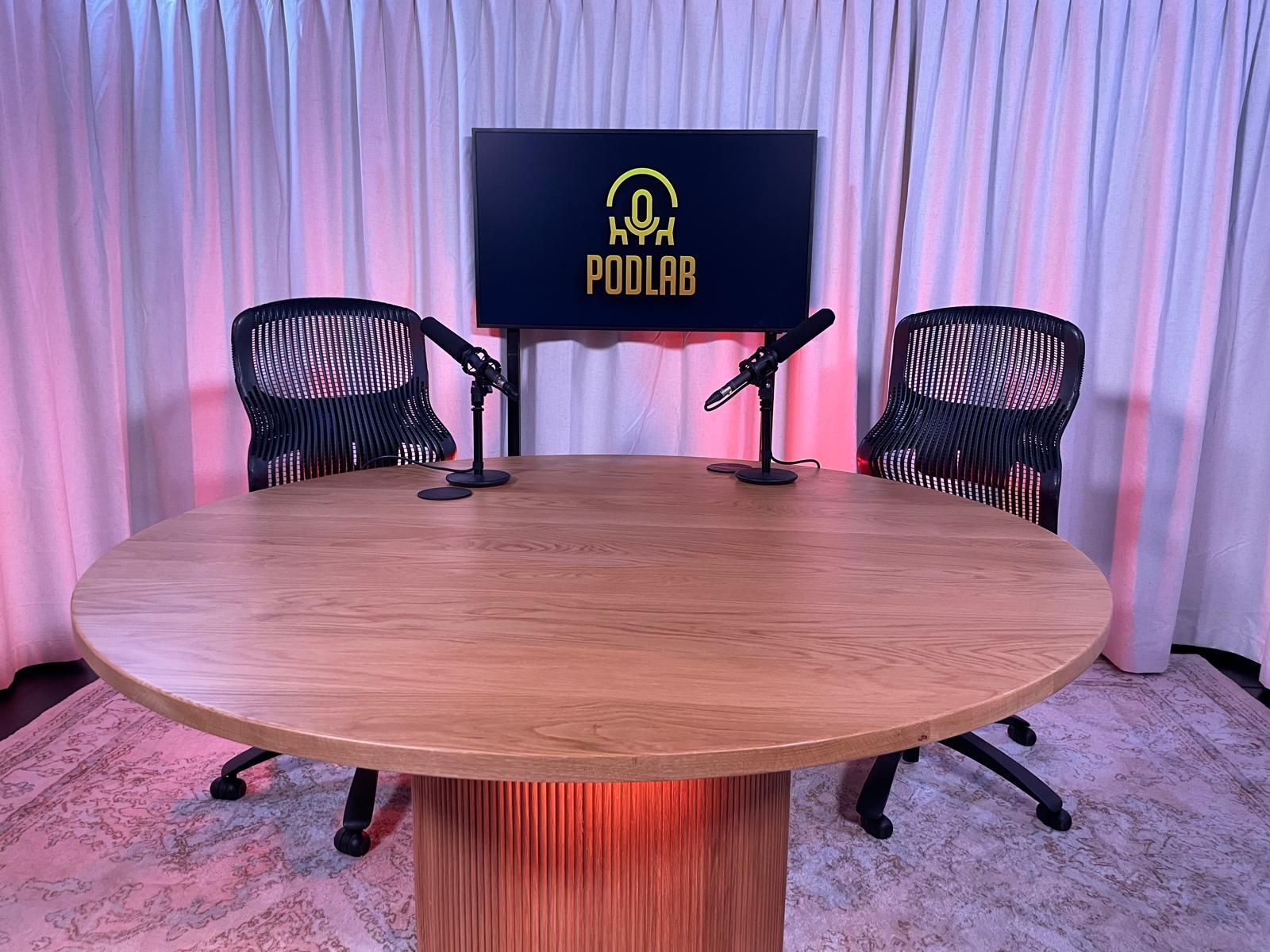 Podcast studio with round wooden table, two black chairs, microphones, and a screen on a pink-lit backdrop