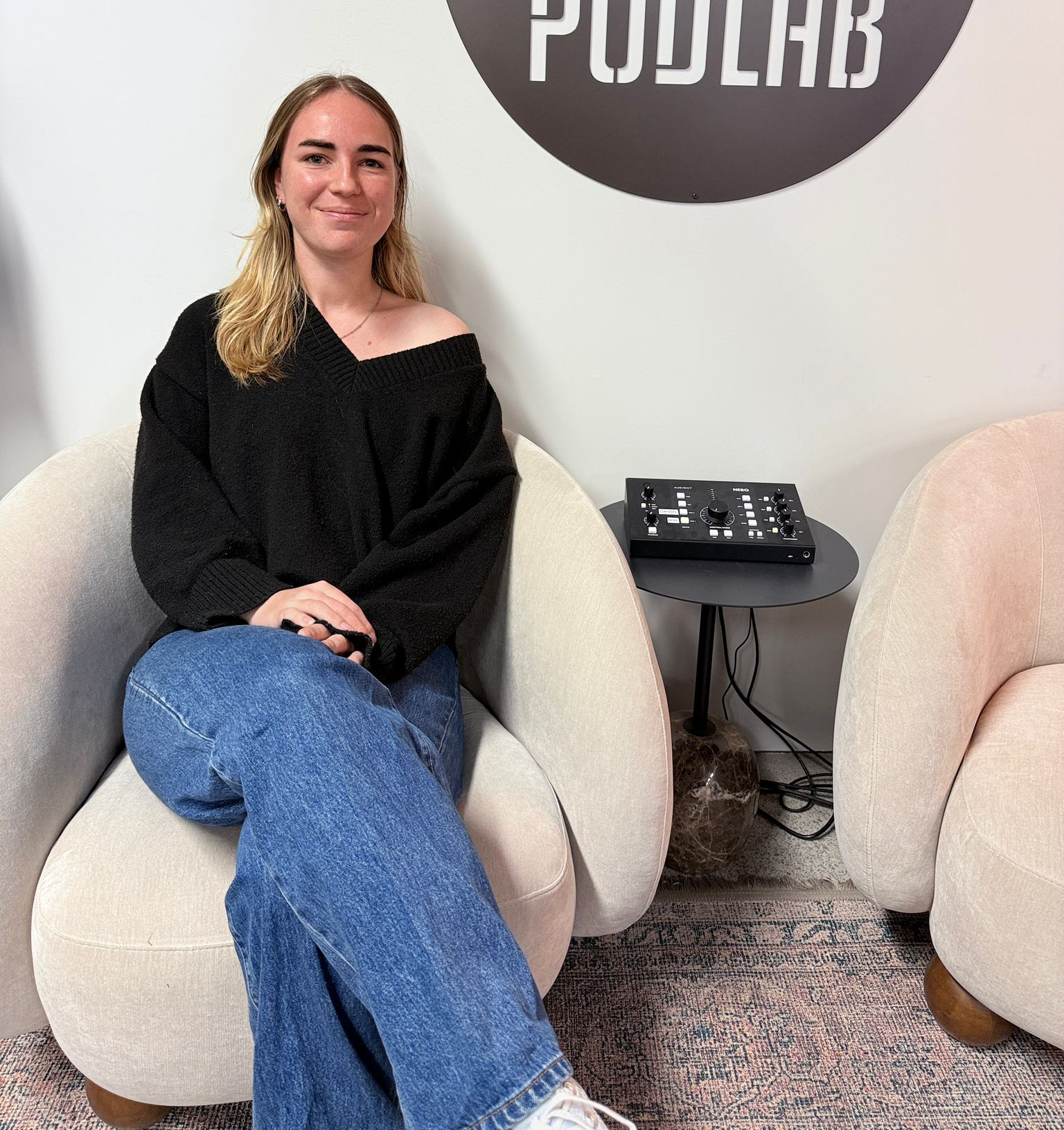 Woman sitting in a cream chair in a PODLAB waiting area, smiling beside a wall logo and phone