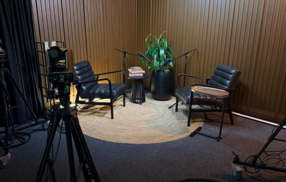 Studio interview set with two black chairs, small table, potted plant, and lighting equipment on a carpeted floor.